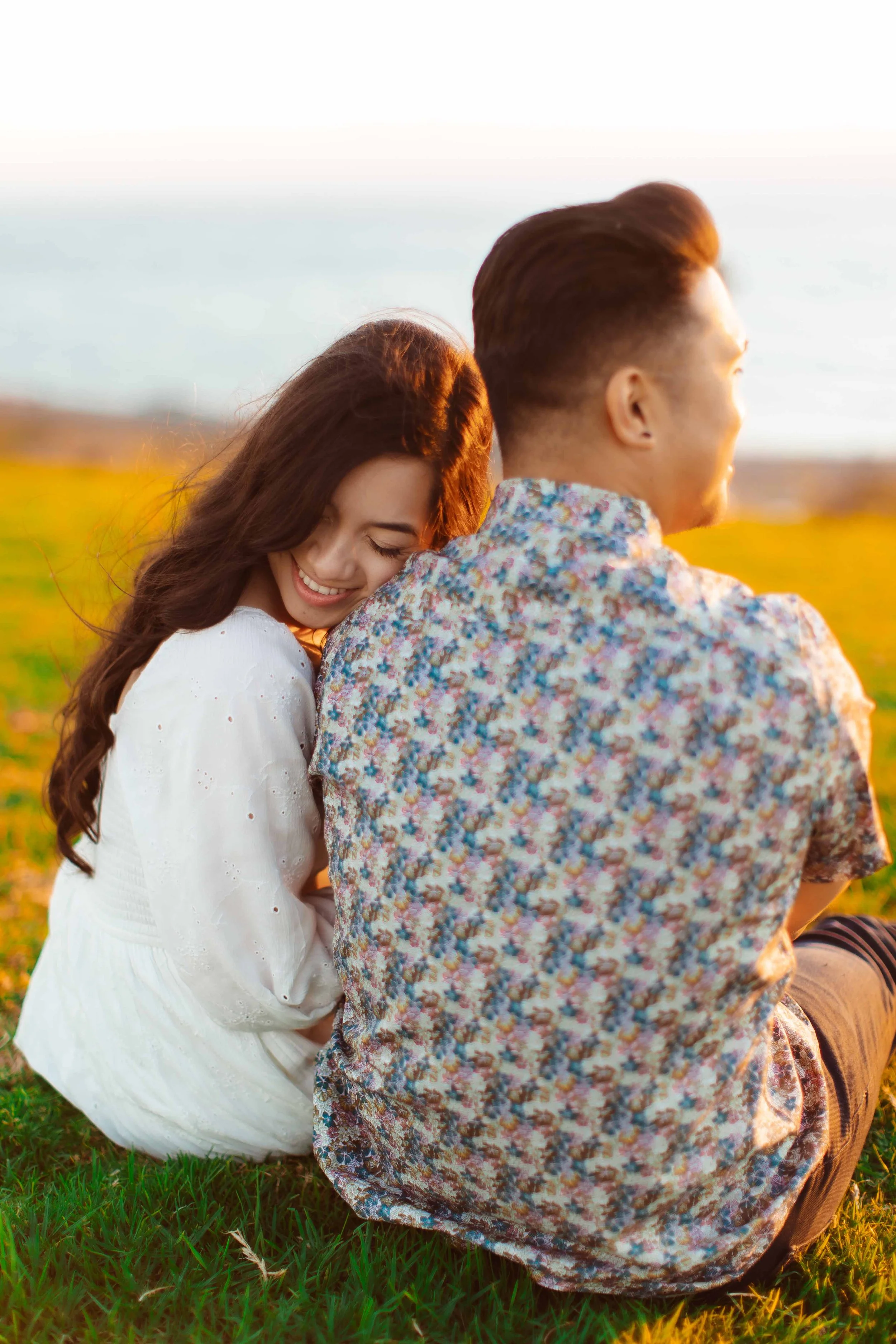 A young woman and man sitting on grass, smiling, with a scenic background of water and open sky during sunset.