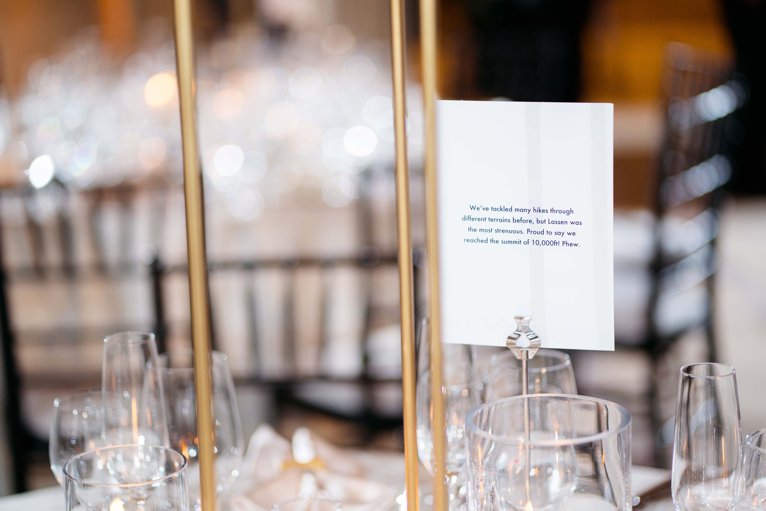 A dining table with glasses and a standing cardholder holding a white card with blue text in a blurred elegant restaurant setting.