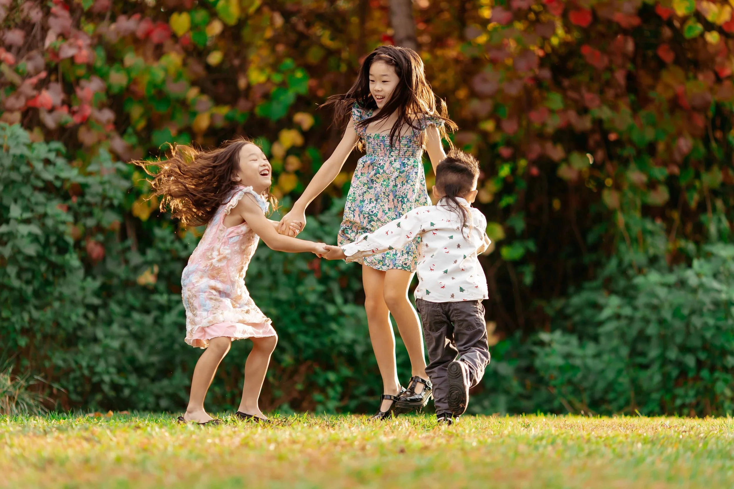 Three children, two girls and a boy, playing together outdoors on grass, holding hands, and smiling, with colorful bushes in the background.