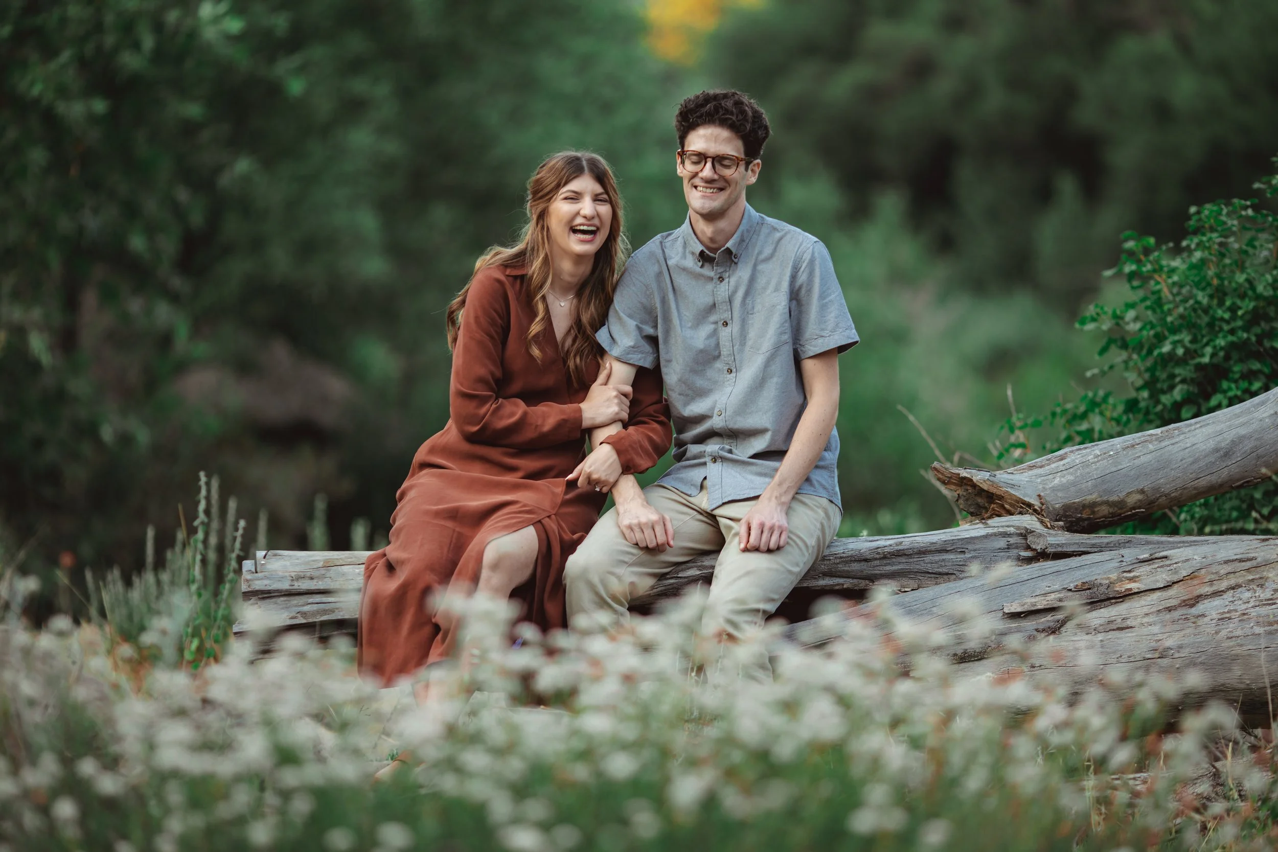 A young couple sitting on a fallen tree in a forest, laughing and enjoying each other's company.