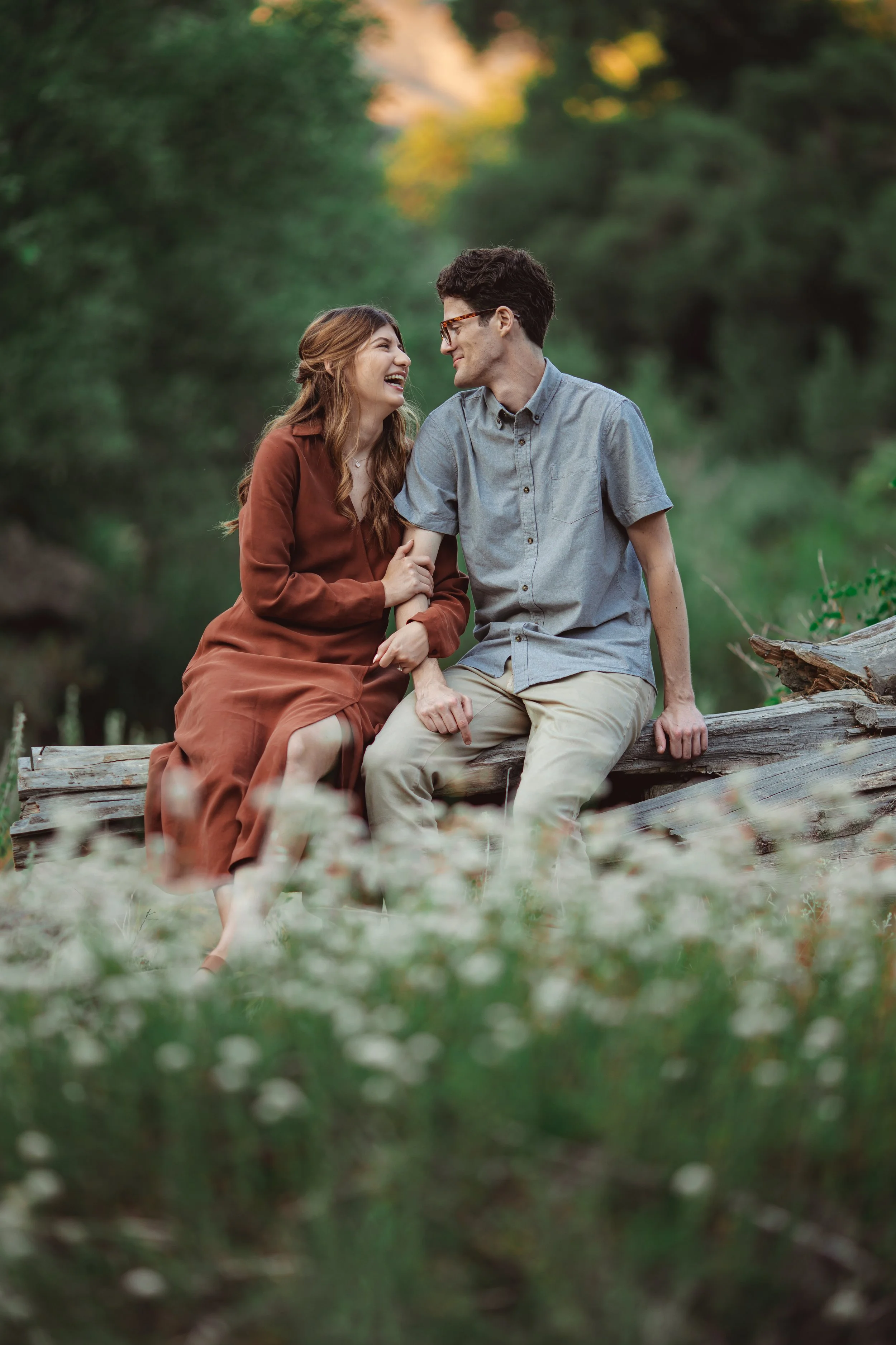 A young couple happily sitting on a fallen log in a lush green outdoor setting, sharing a moment of laughter and affection.