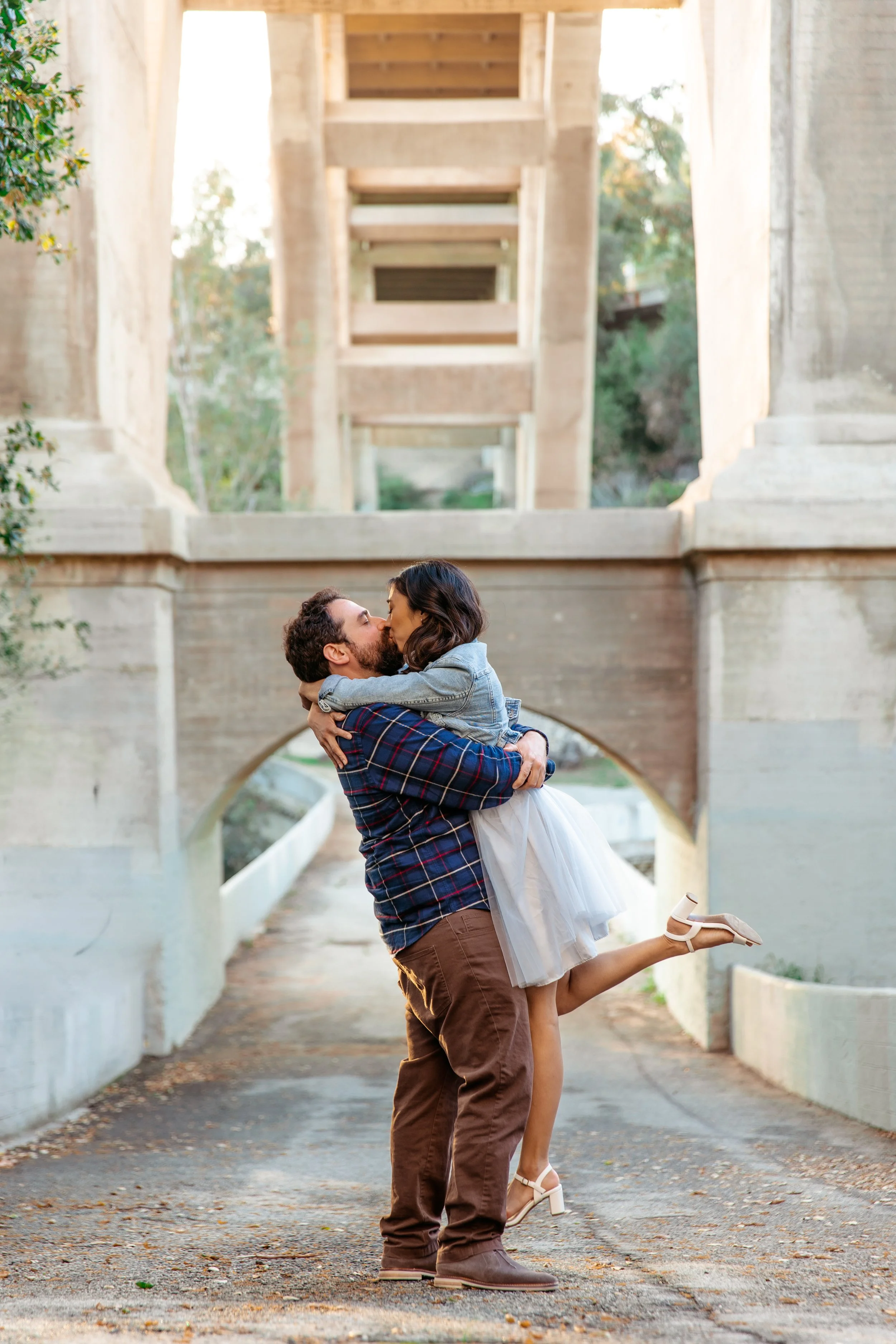 A man lifting a woman and kissing under a bridge in a park during daylight.