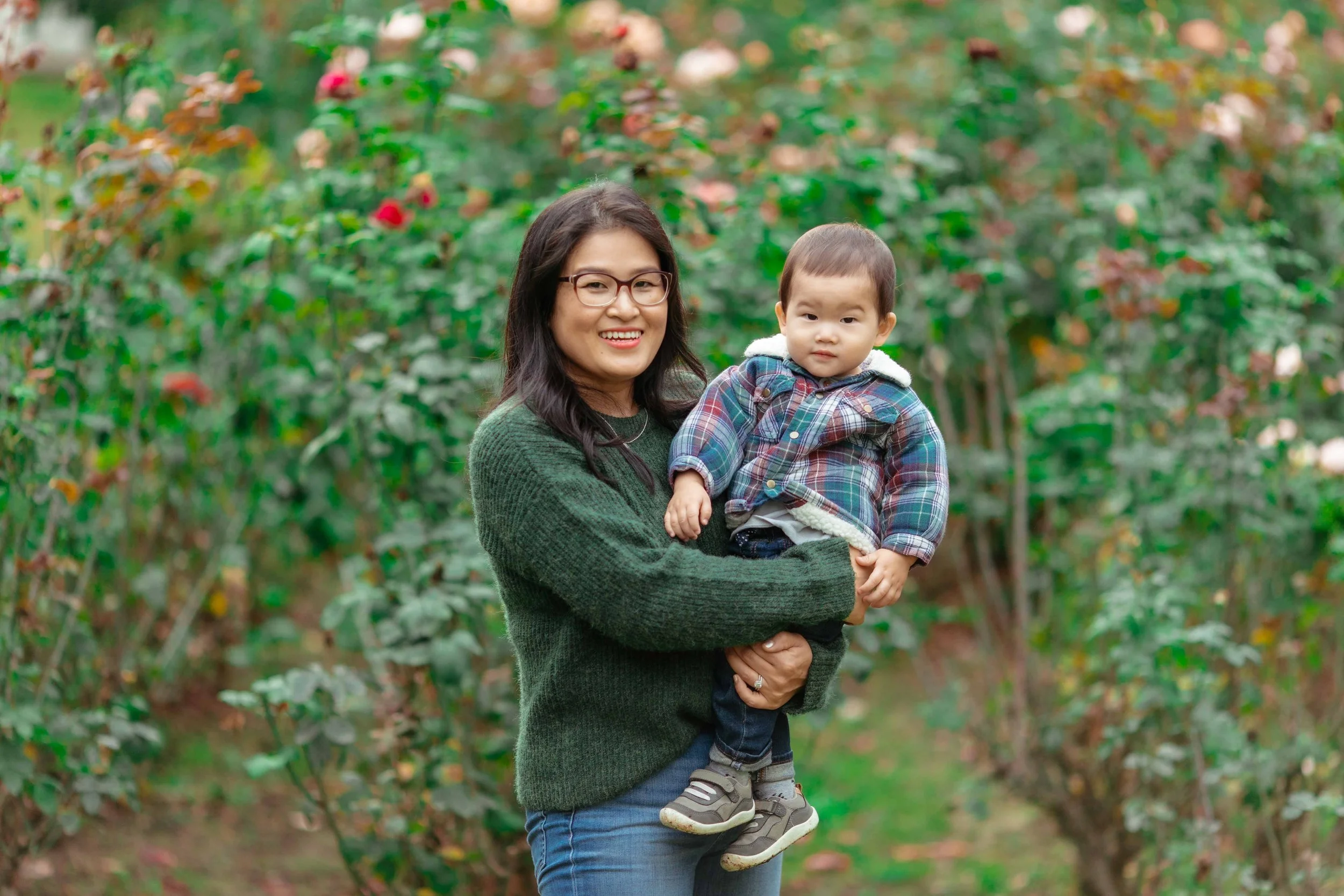 A woman smiling and holding a young boy in a garden with pink and white flowers and green foliage.