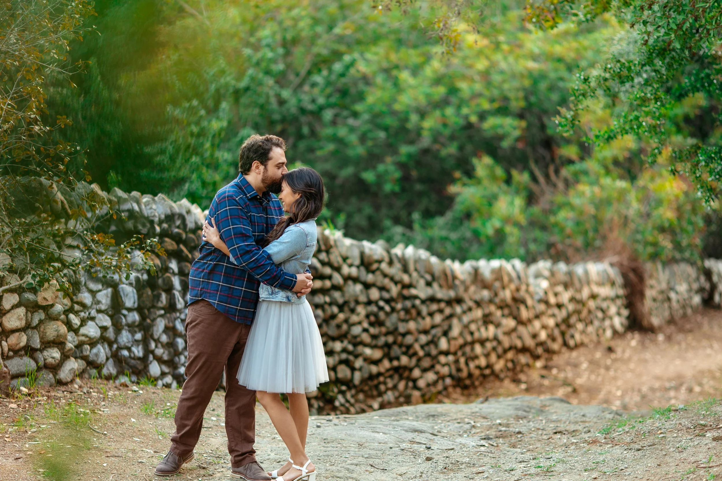 A couple hugging on a dirt path outdoors, with a stone wall and green trees in the background.