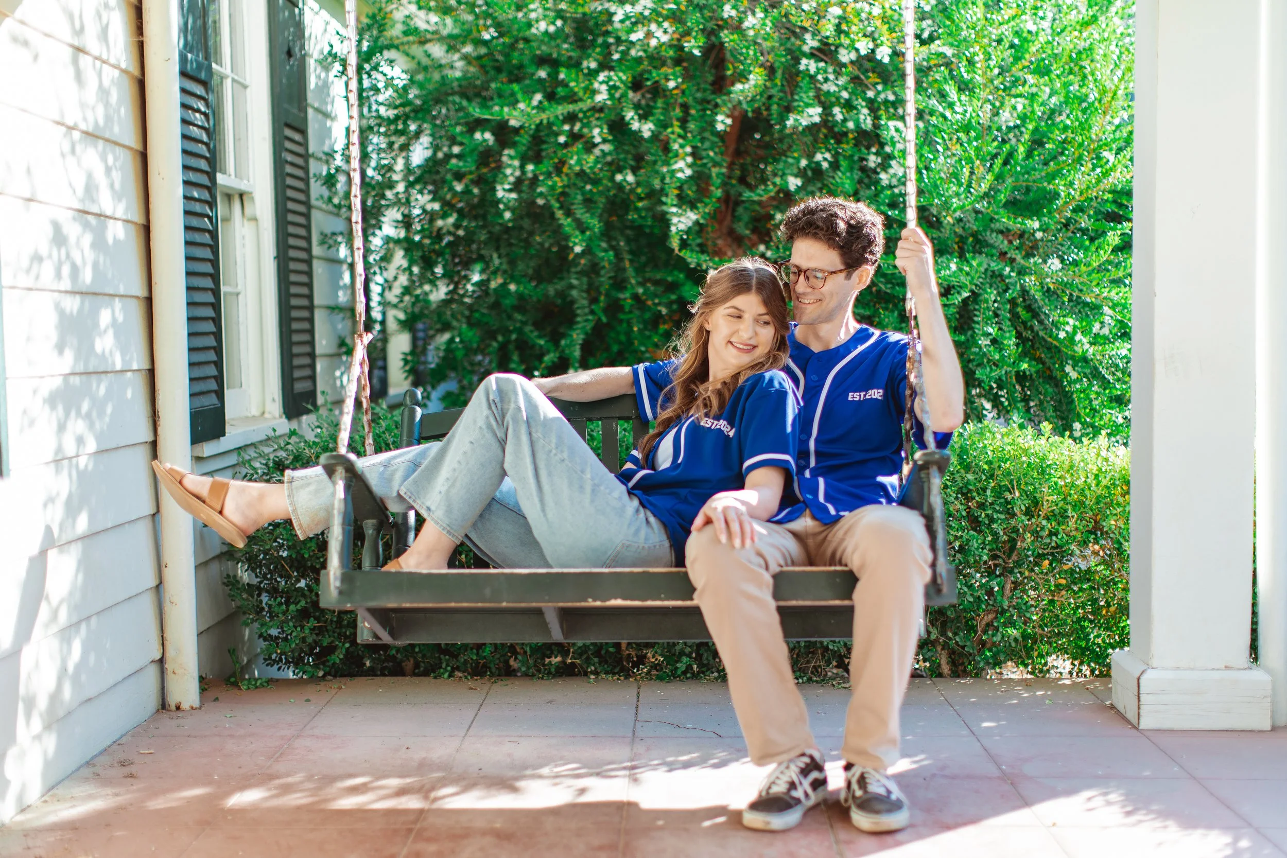 A young couple wearing matching blue jerseys sitting on a porch swing, smiling and enjoying each other's company. The porch has a white house with black shutters and lush greenery in the background.