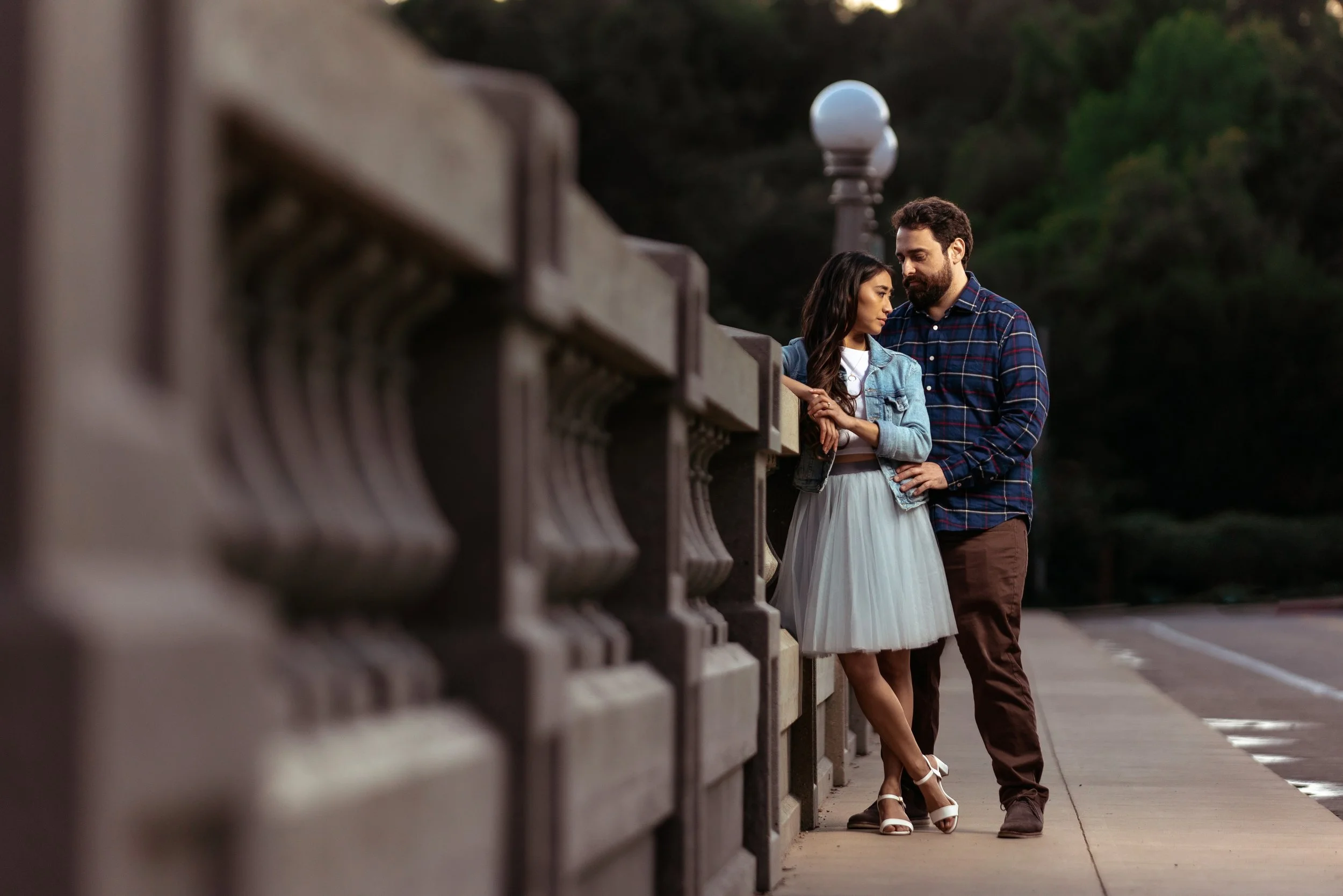 A young woman and a man standing close together on a bridge, engaged in a tender moment, with trees in the background.
