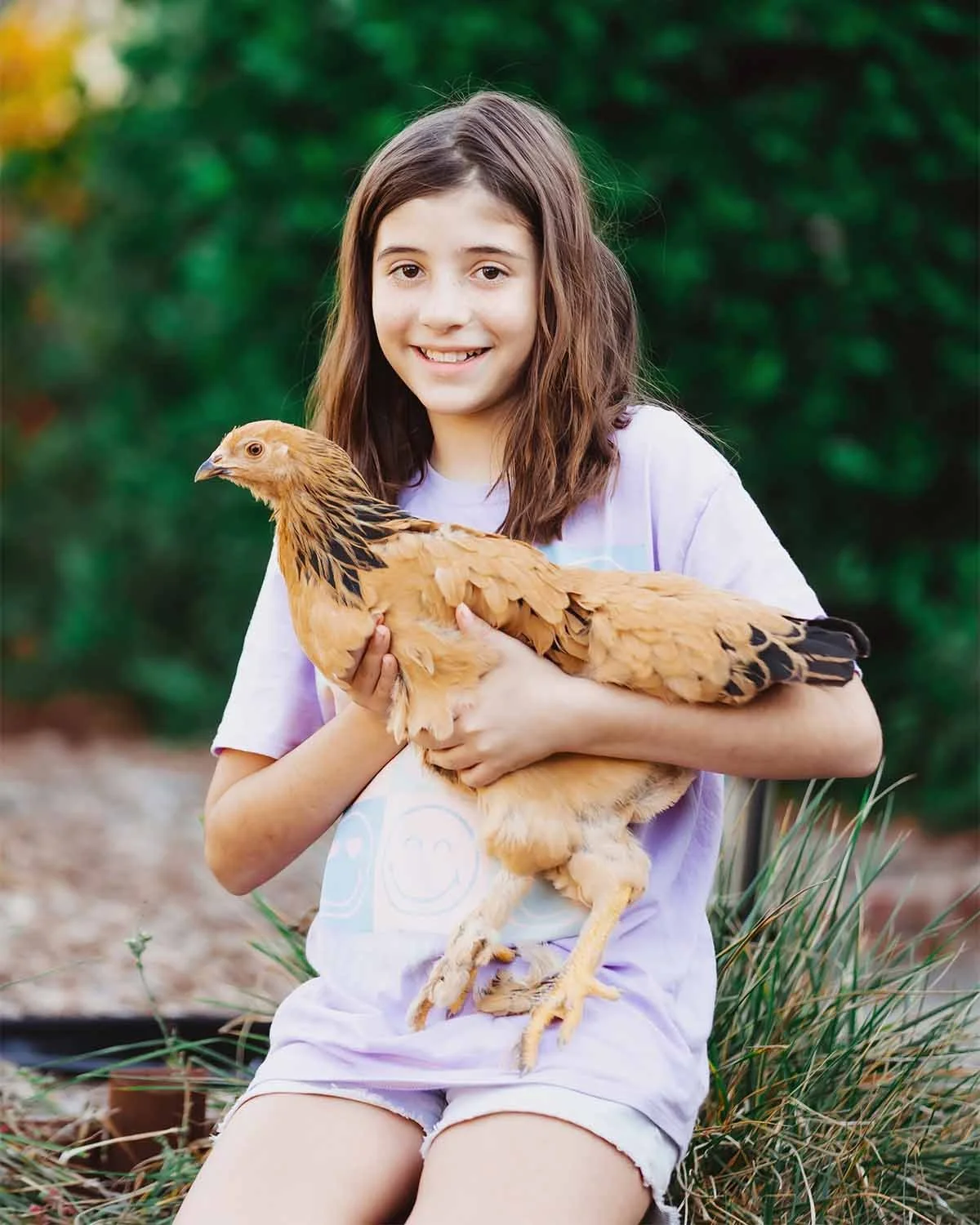 A young girl sitting outdoors holding a chicken, with greenery in the background.
