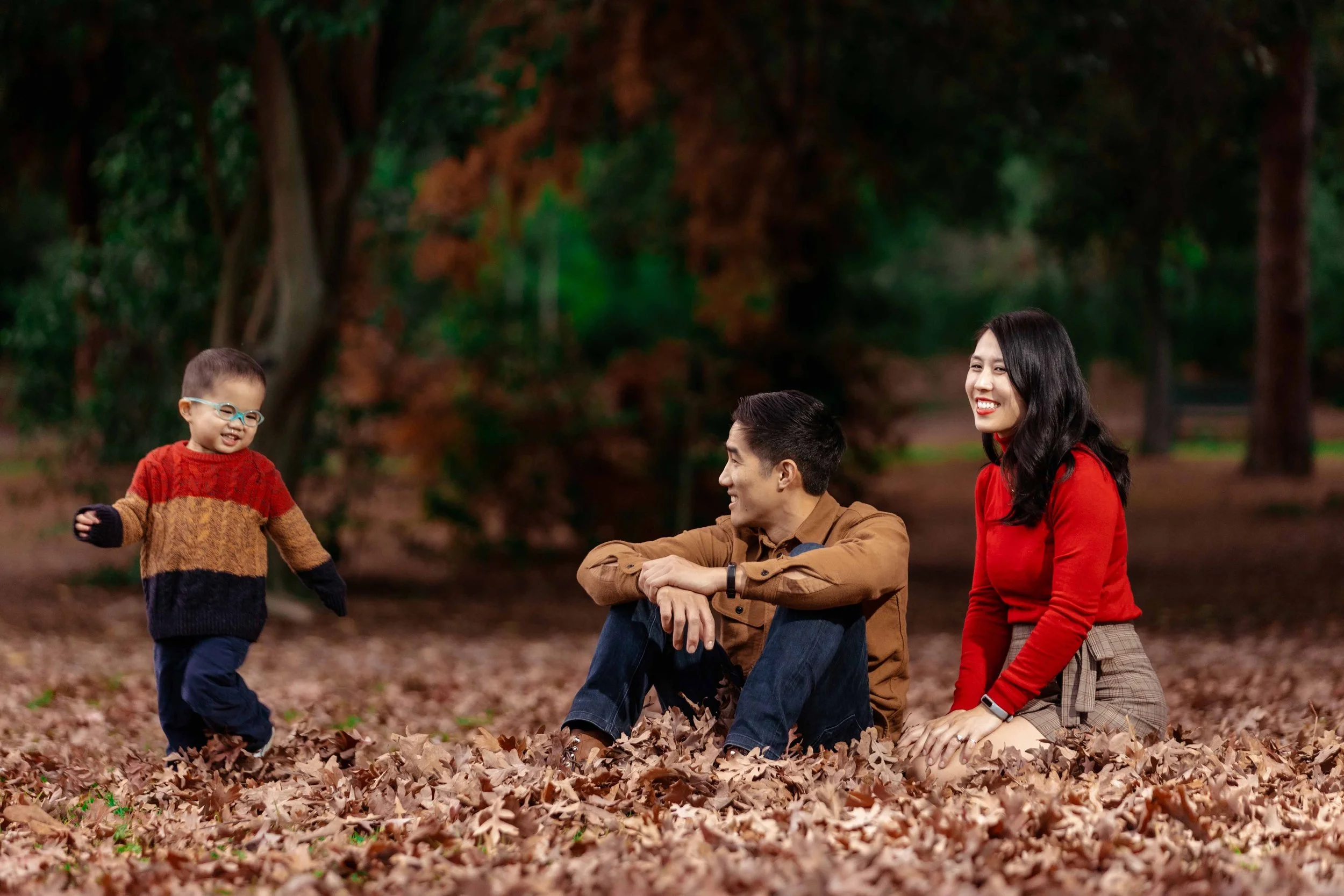 A happy family of three outdoors during autumn, with the father and mother sitting on the fallen leaves while their young son, wearing glasses and a colorful sweater, stands and smiles at them.