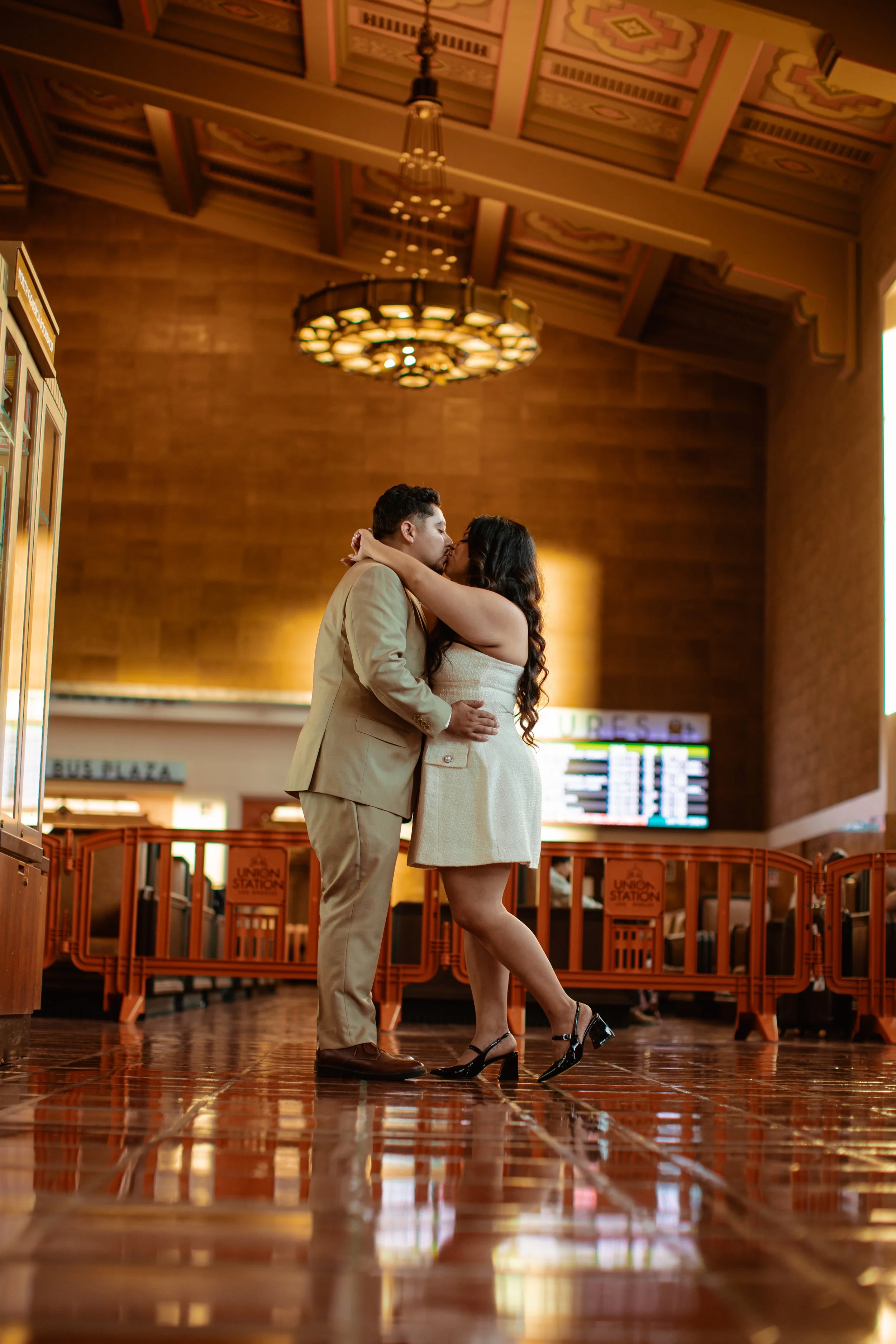 A couple sharing a kiss at a train station with a high ceiling and decorative lighting.
