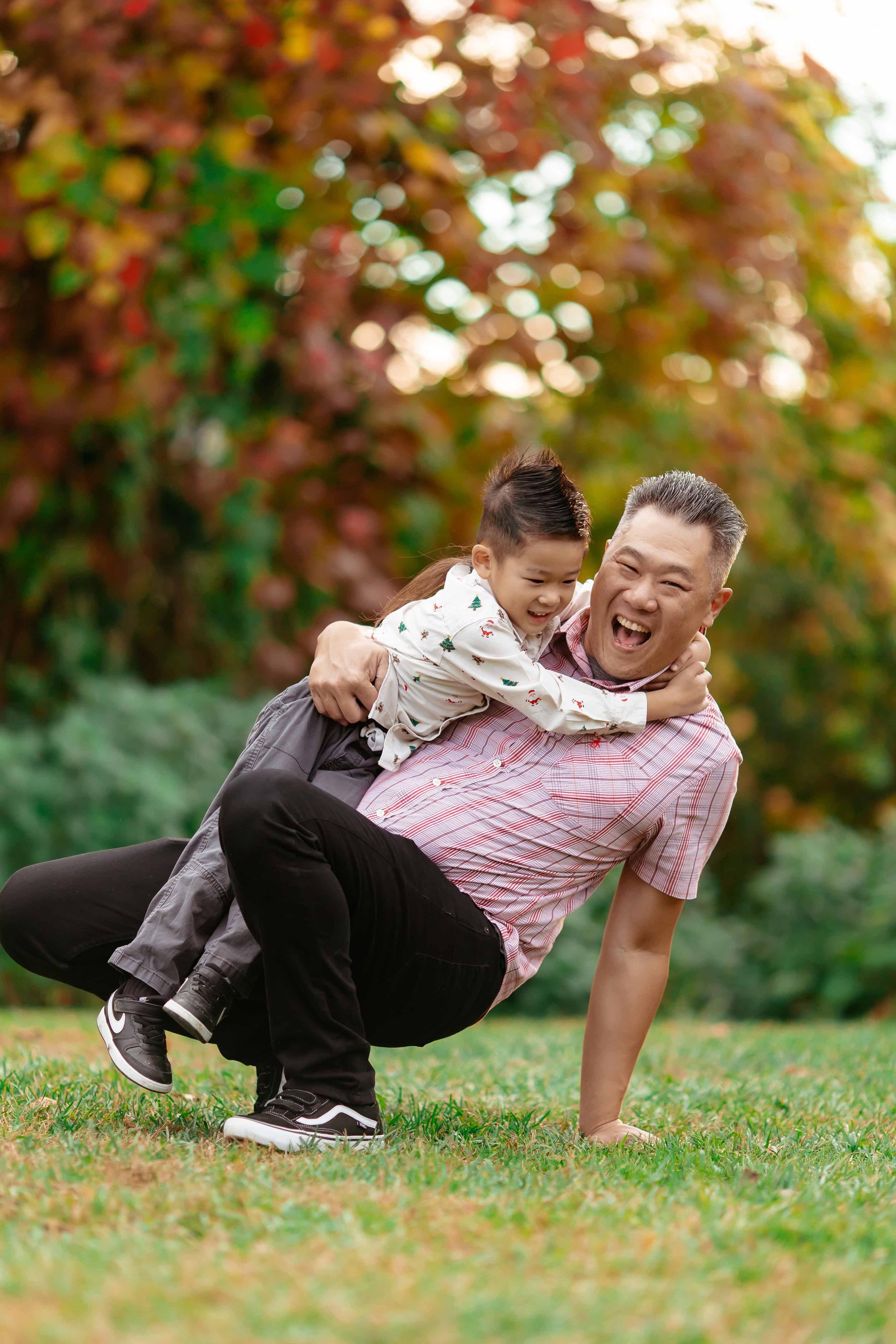 A man and a young girl playing outdoors on grass, with trees and colorful foliage in the background, both smiling and having fun.