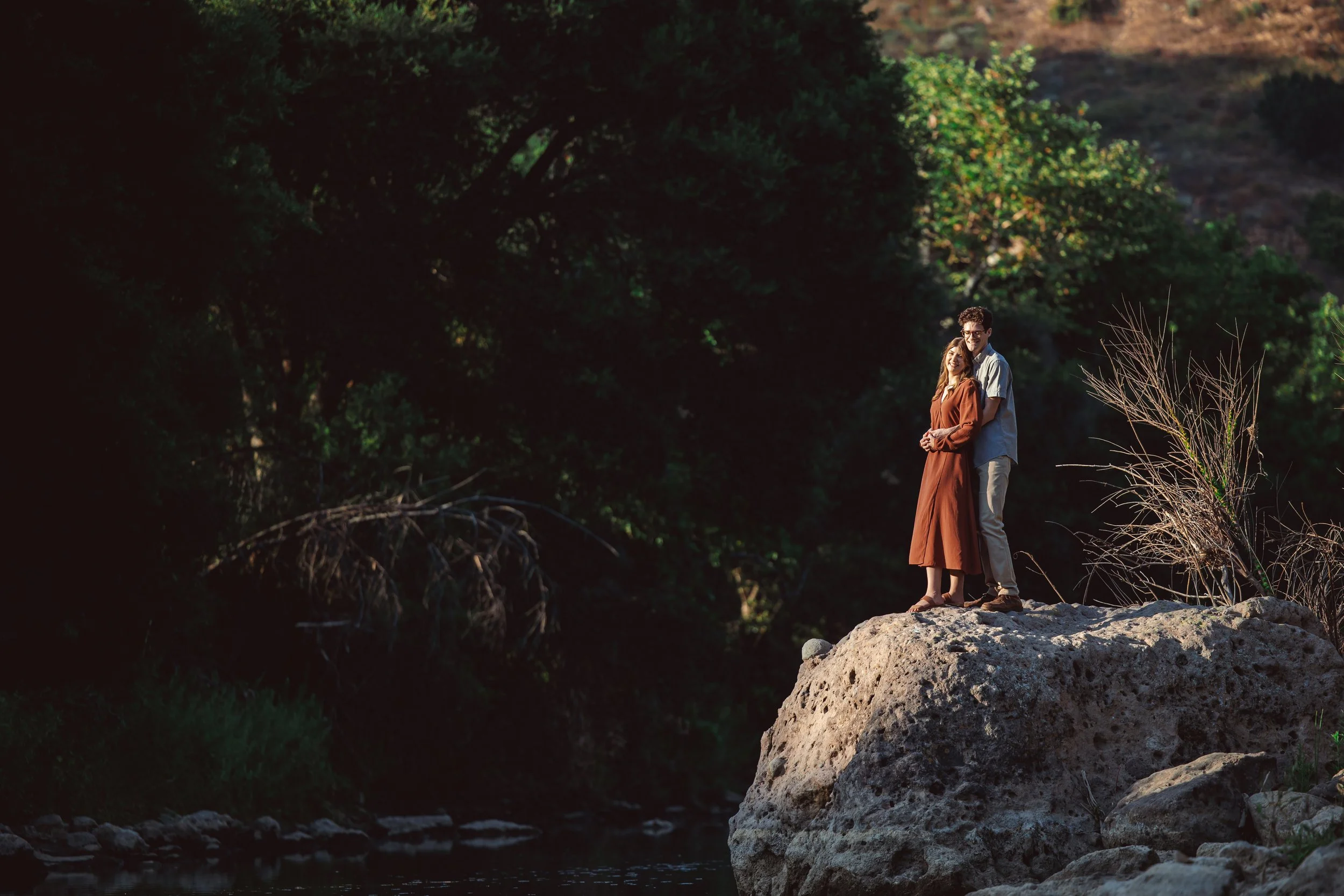 A young couple stands on a large rock beside a river, embracing and smiling in a natural, wooded outdoor setting.