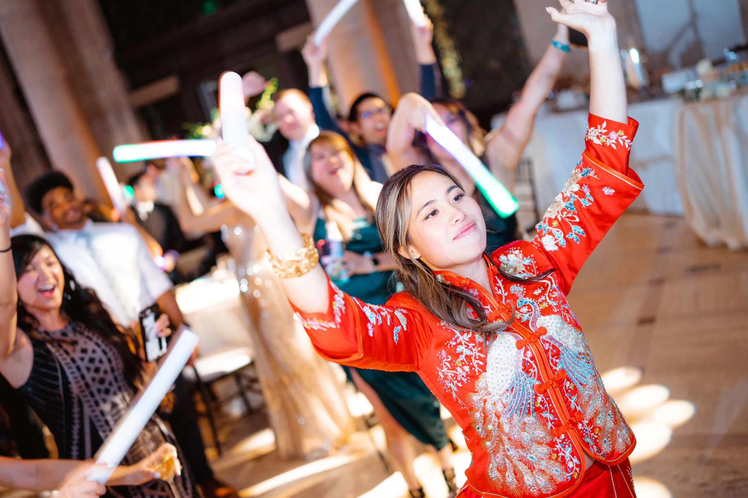 A woman in a traditional Chinese red dress dancing at a celebration, surrounded by friends who are also dancing and celebrating indoors.