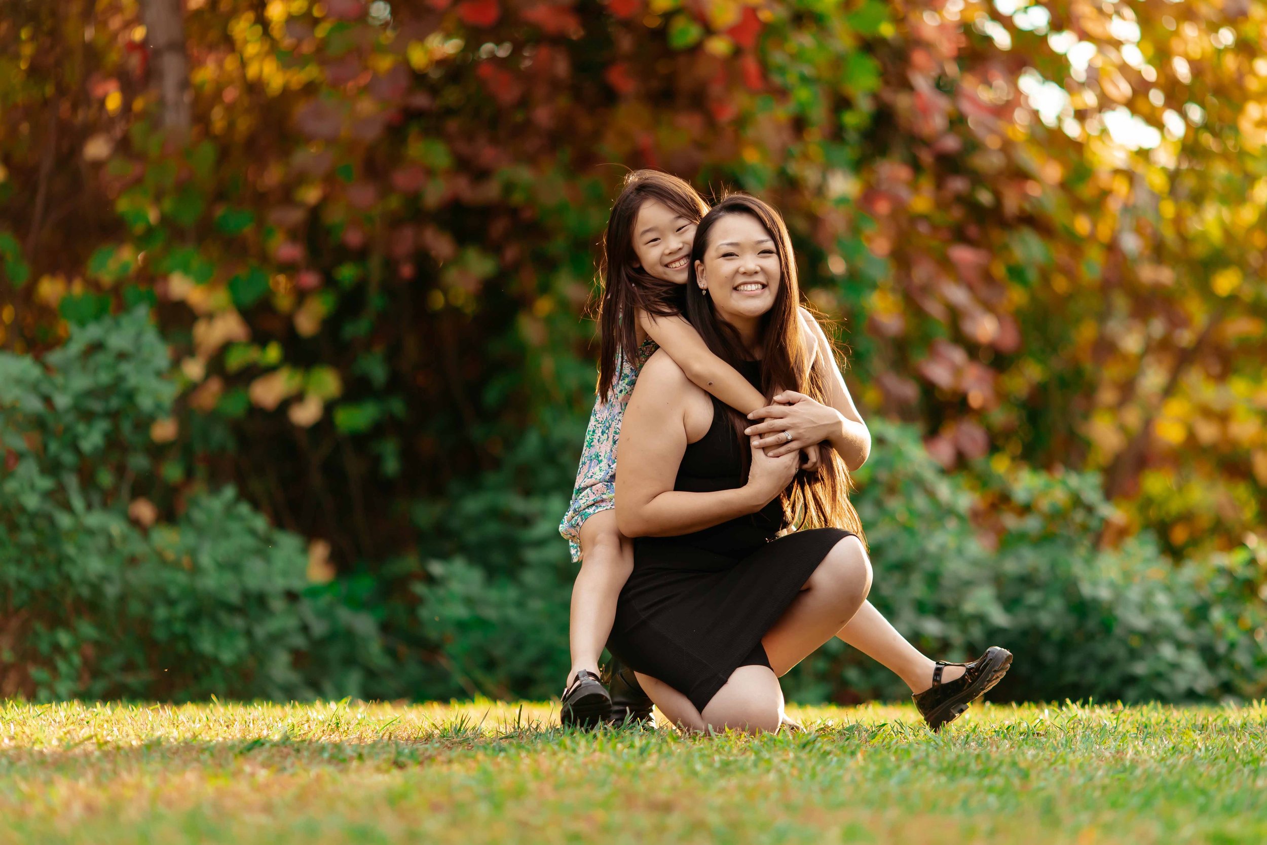 A woman kneeling on grass and holding a young girl on her back, both smiling in a park with autumn-colored trees.