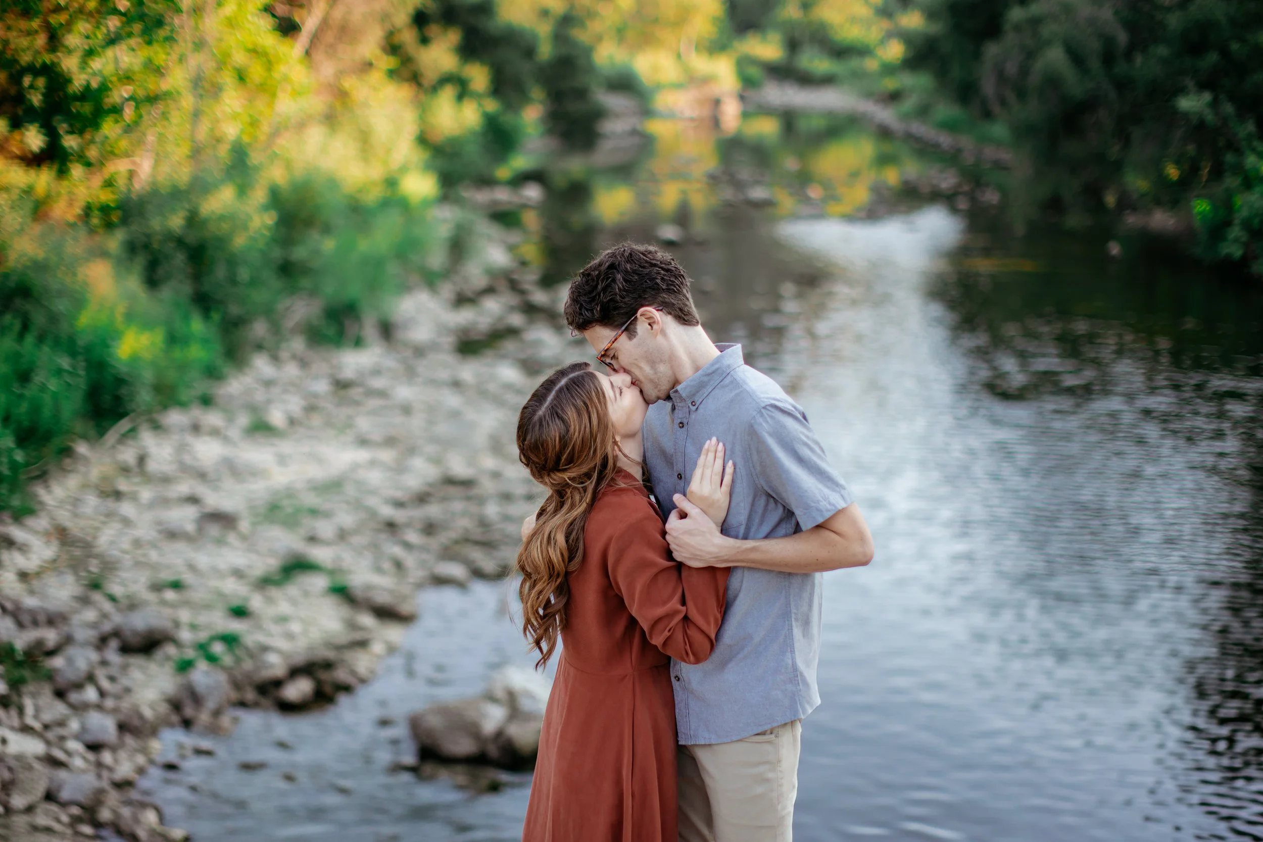 A young couple sharing a kiss by a river in a forested area.