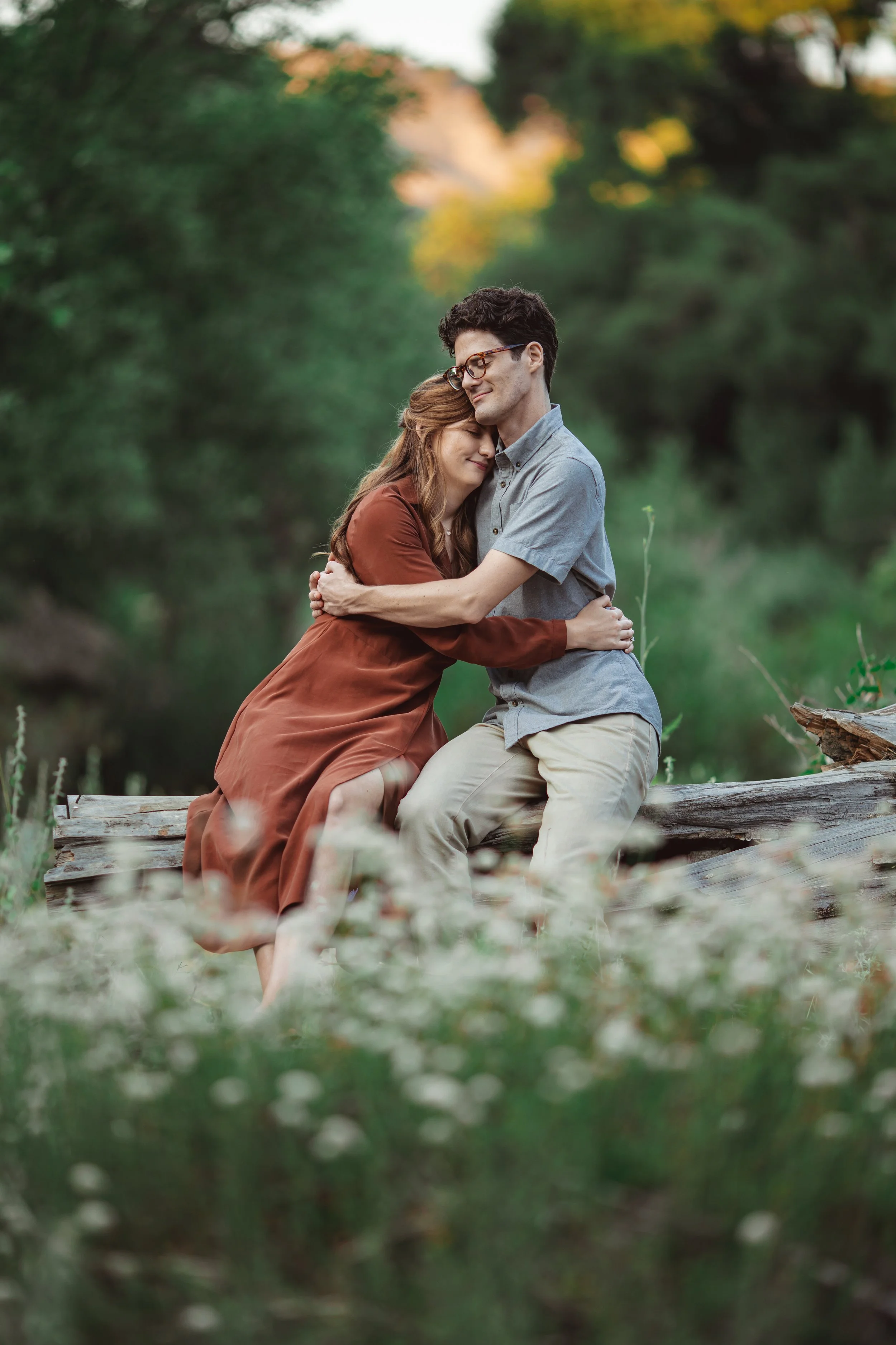 A young couple sitting on a fallen tree, embracing and smiling in a lush, green outdoor setting with soft focus foliage in the background.