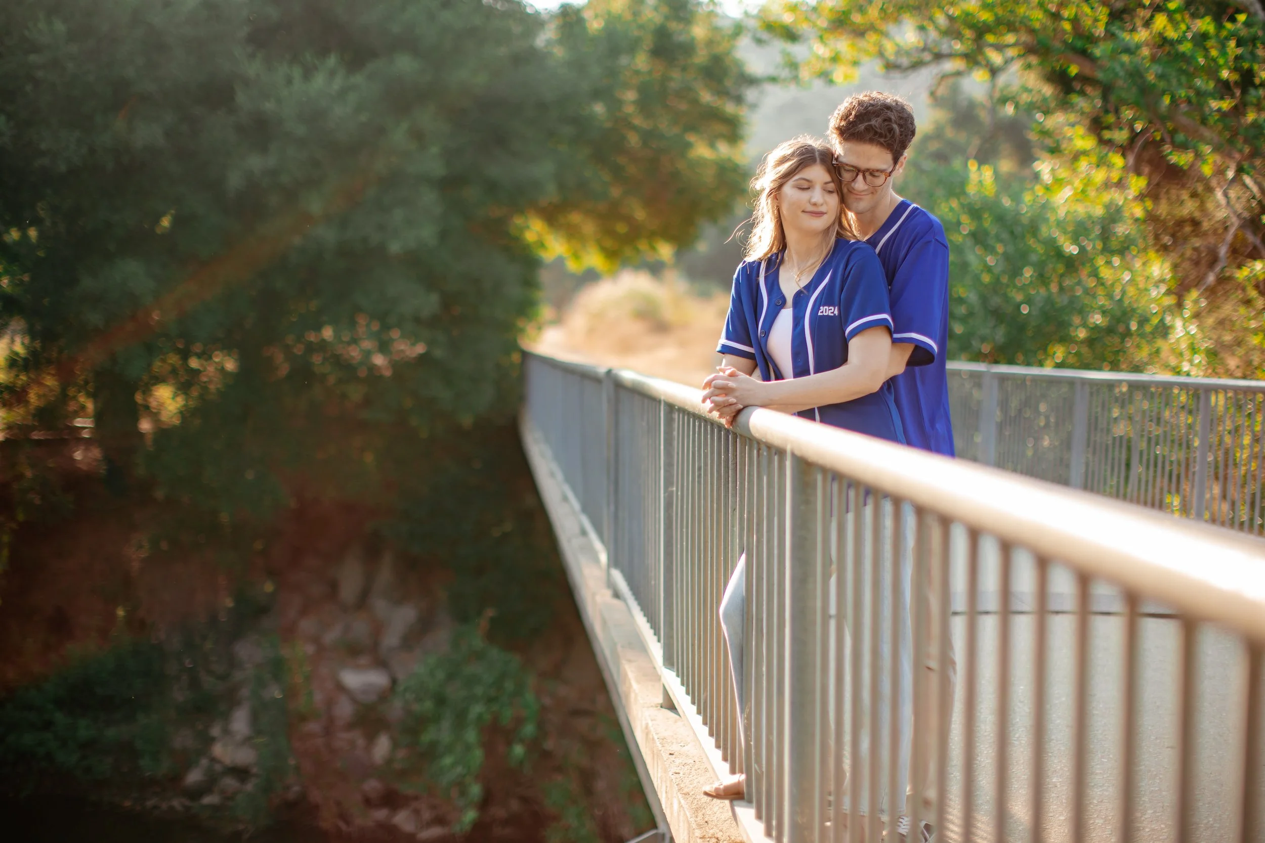 A young couple dressed in matching blue sports jerseys embraces on a bridge, overlooking a canyon with lush green trees and rocky terrain.