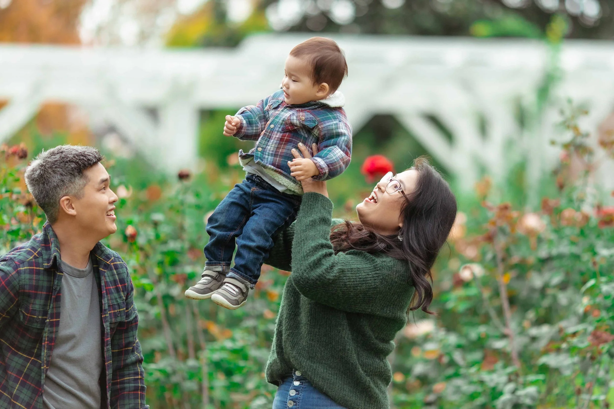 A family of three enjoying time outdoors in a garden or park, with a woman lifting a young boy while a man watches and smiles.