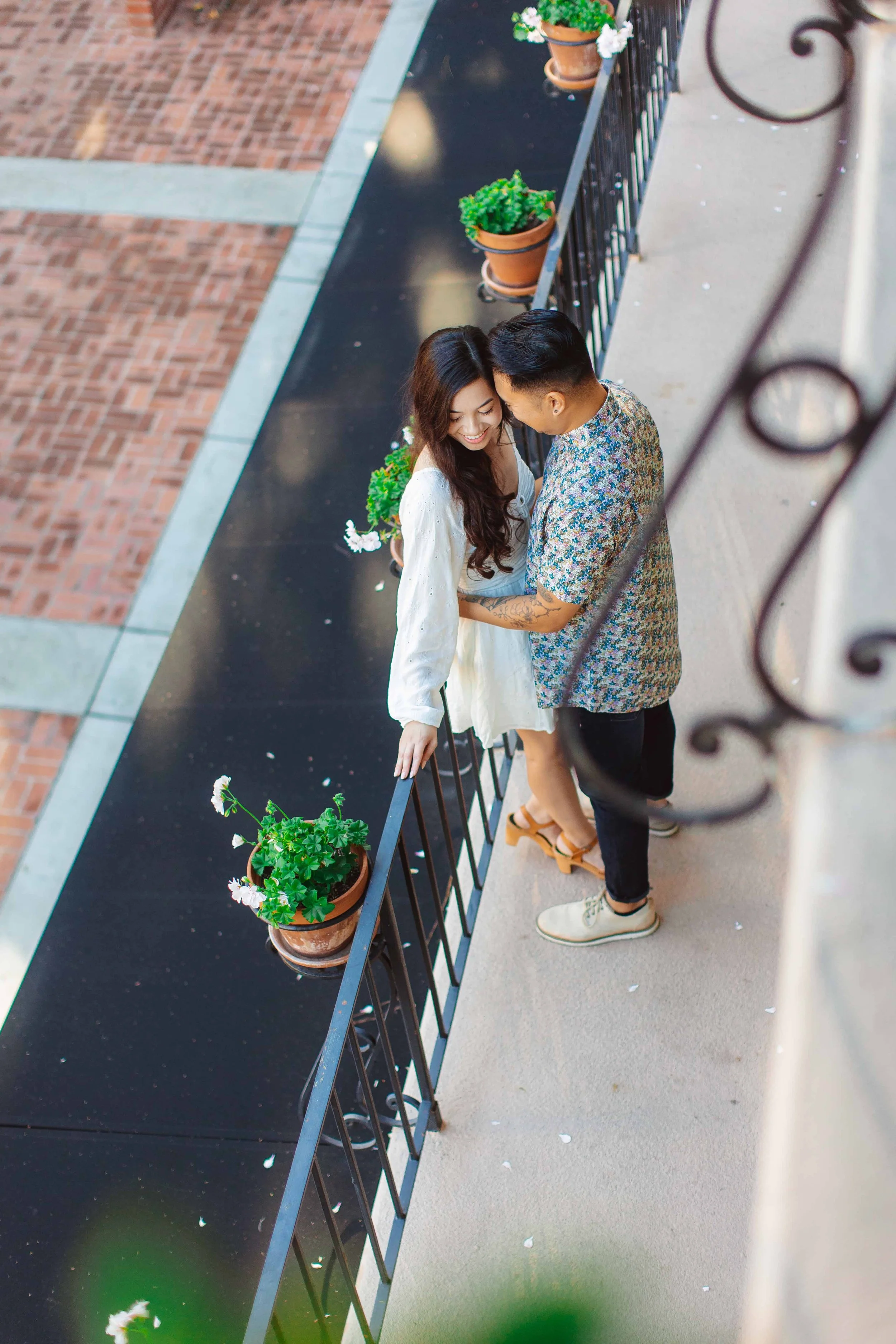 A woman and a man on a narrow balcony are smiling and embracing each other. The woman is wearing a white dress, and the man is wearing a patterned shirt. There are potted plants with flowers on the balcony.