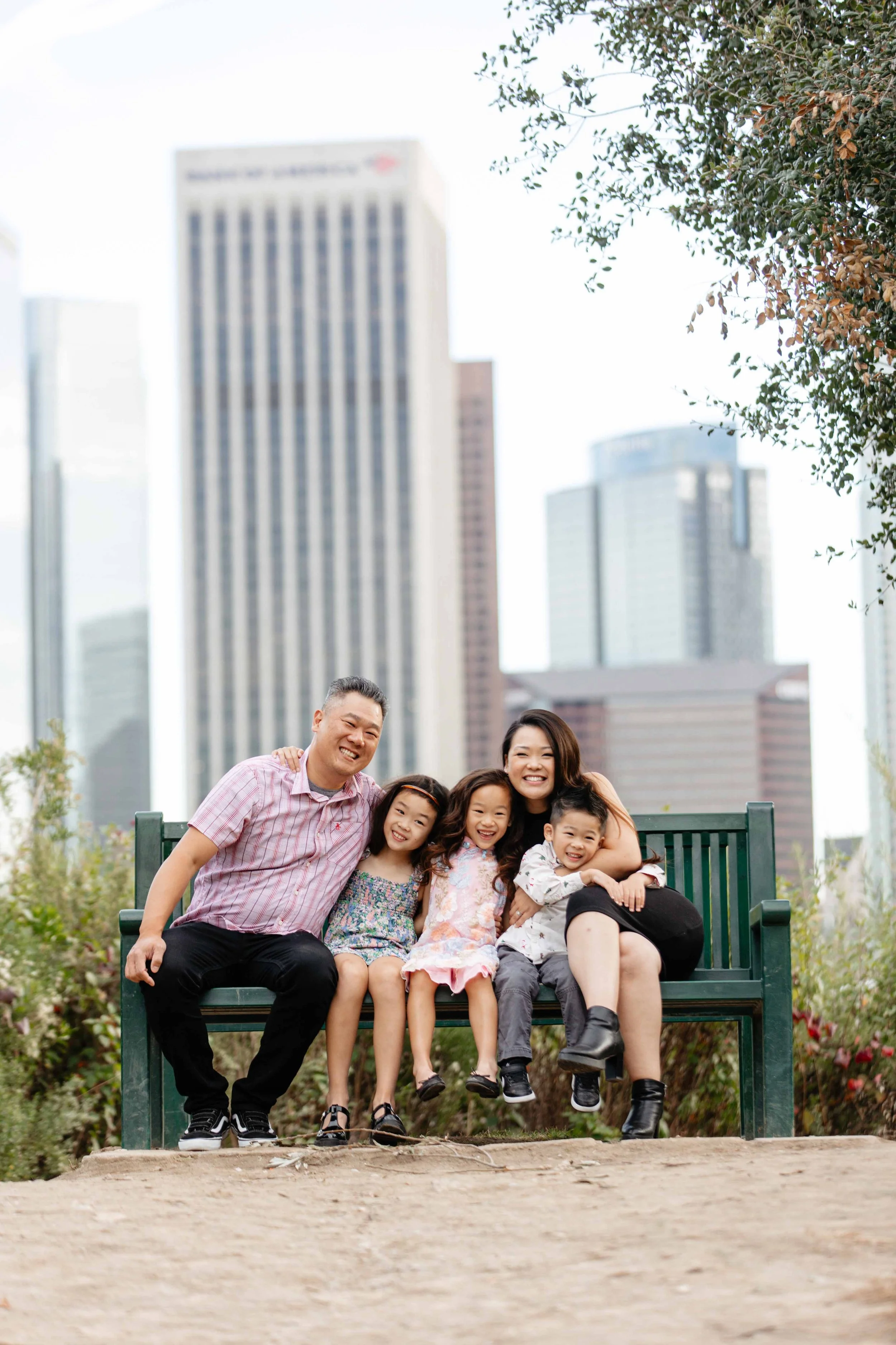 A happy family of five sitting on a park bench in front of a city skyline with tall buildings, smiling and enjoying a sunny day.