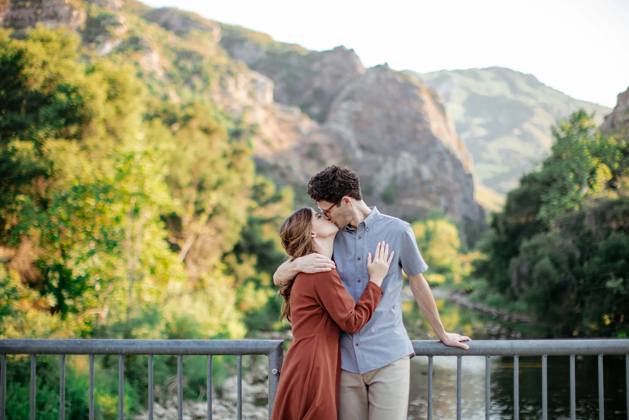 A couple sharing a kiss on a bridge with a scenic mountain and green landscape in the background.
