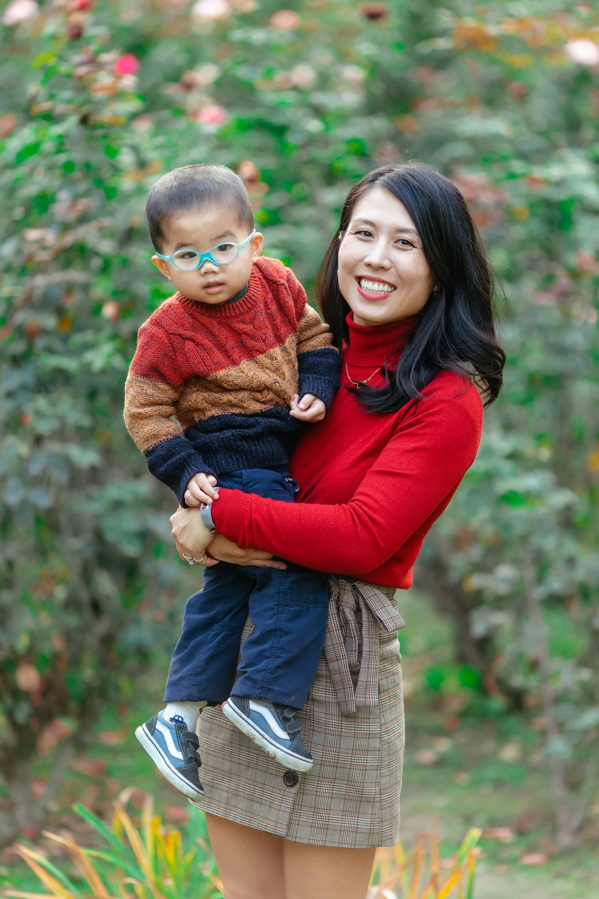 A smiling woman holding a young boy outdoors in a garden or park with green foliage and flowers in the background.