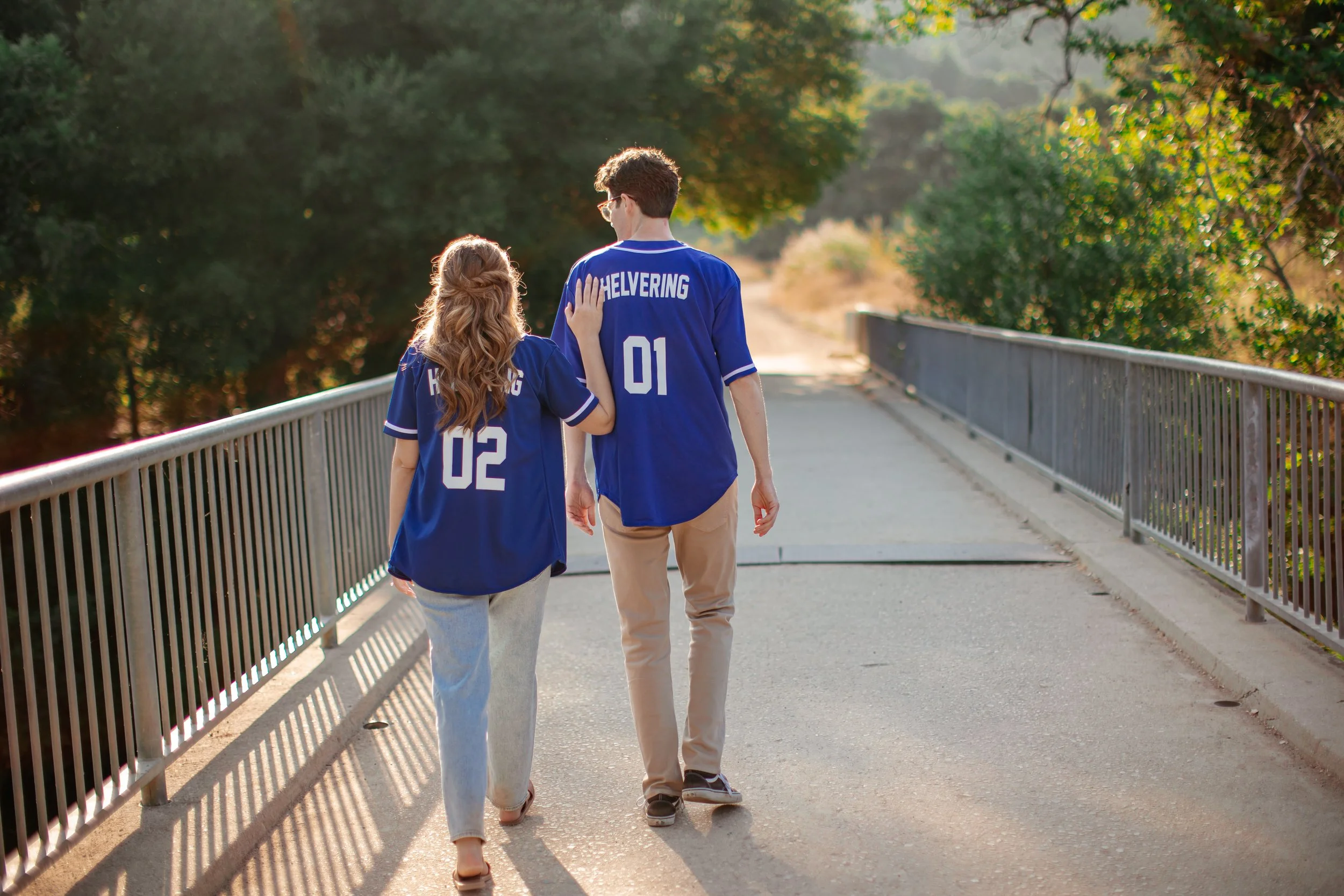 A young couple walking arm in arm on a bridge, wearing matching blue sports jerseys with numbers 02 and 01 and the word "HOVERING" printed on the back, surrounded by greenery and trees.