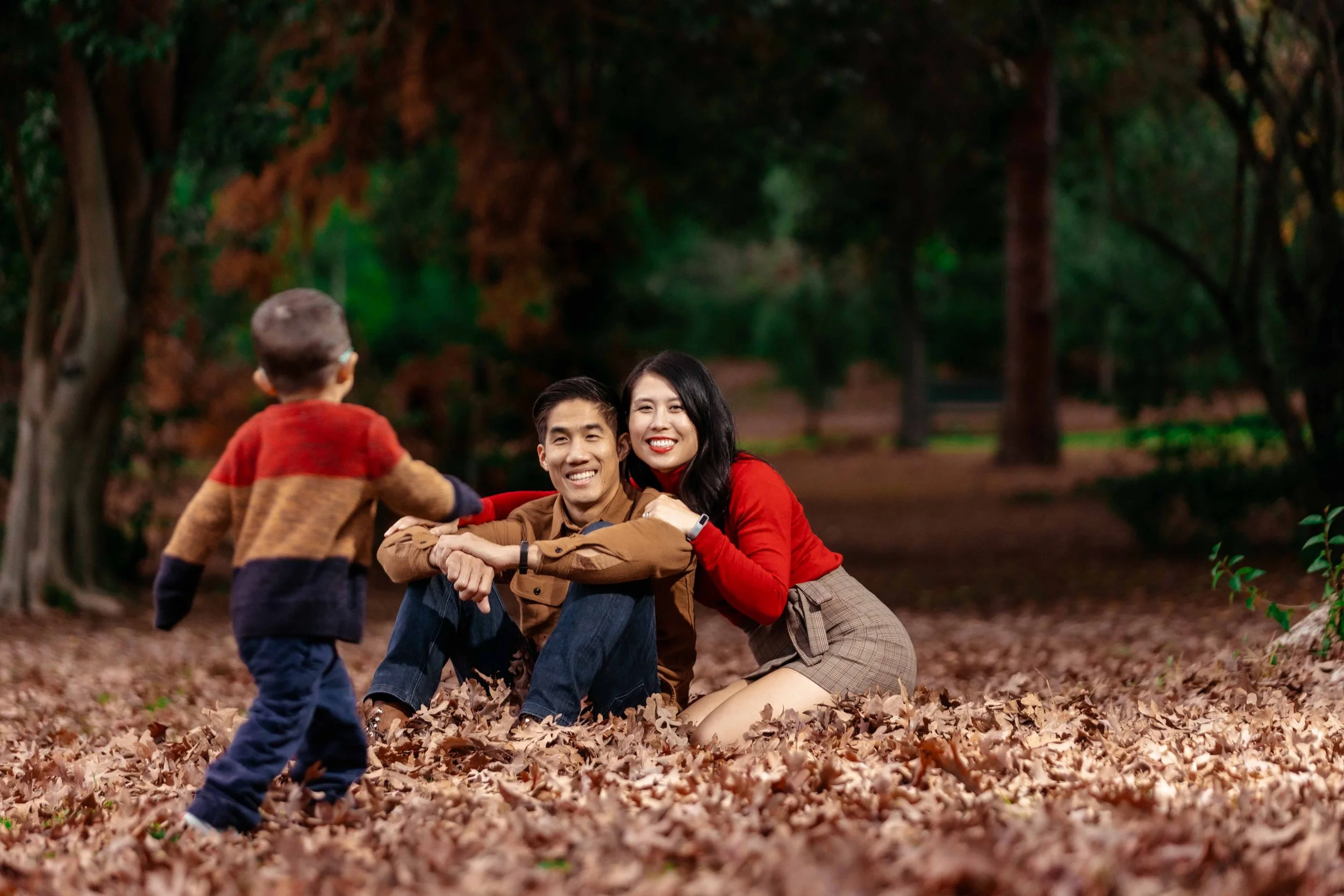 A happy family in a park surrounded by fallen autumn leaves. The mother and father are sitting on the ground, smiling, while their young child approaches them from the front.