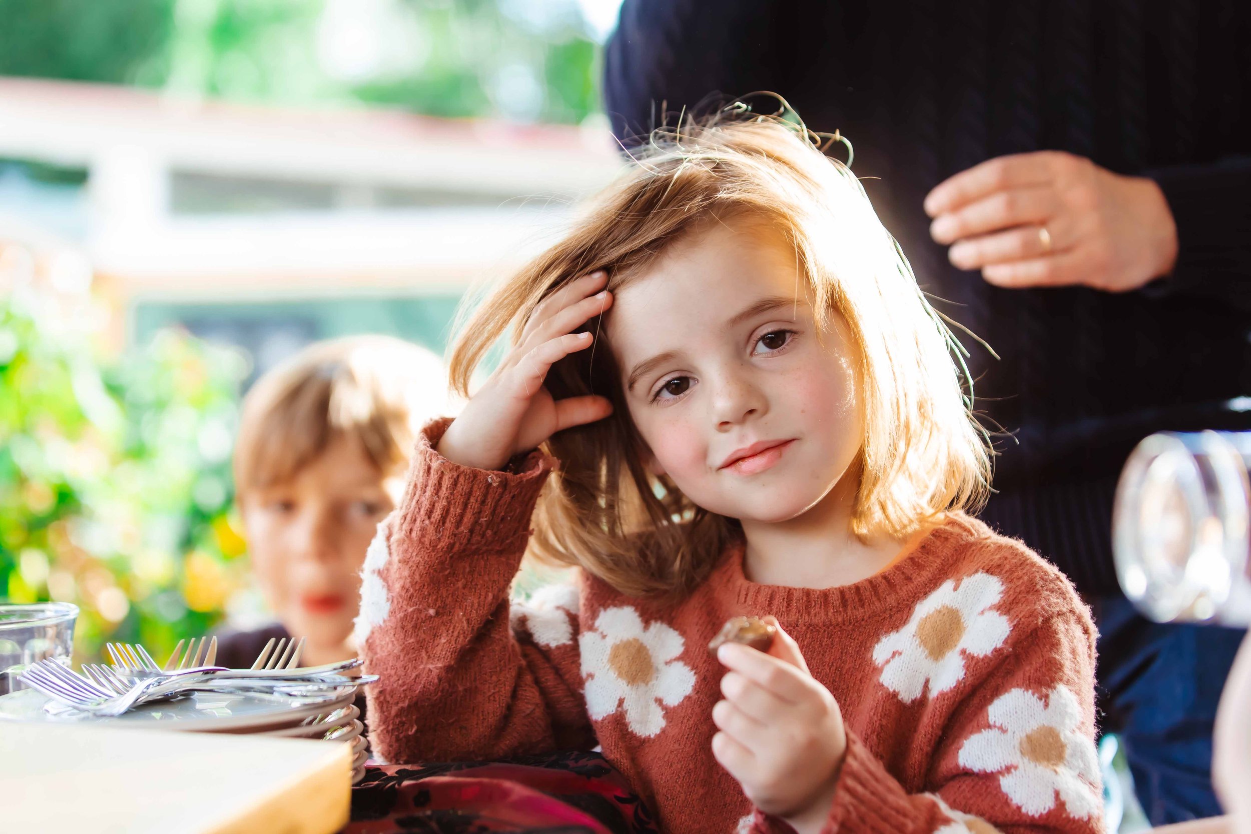 A young girl with light brown hair wearing a brown sweater with white flowers, sitting at a table with a plate of food and holding a piece of food in her hand. She is touching her head with her left hand and looking at the camera with a slight smile.