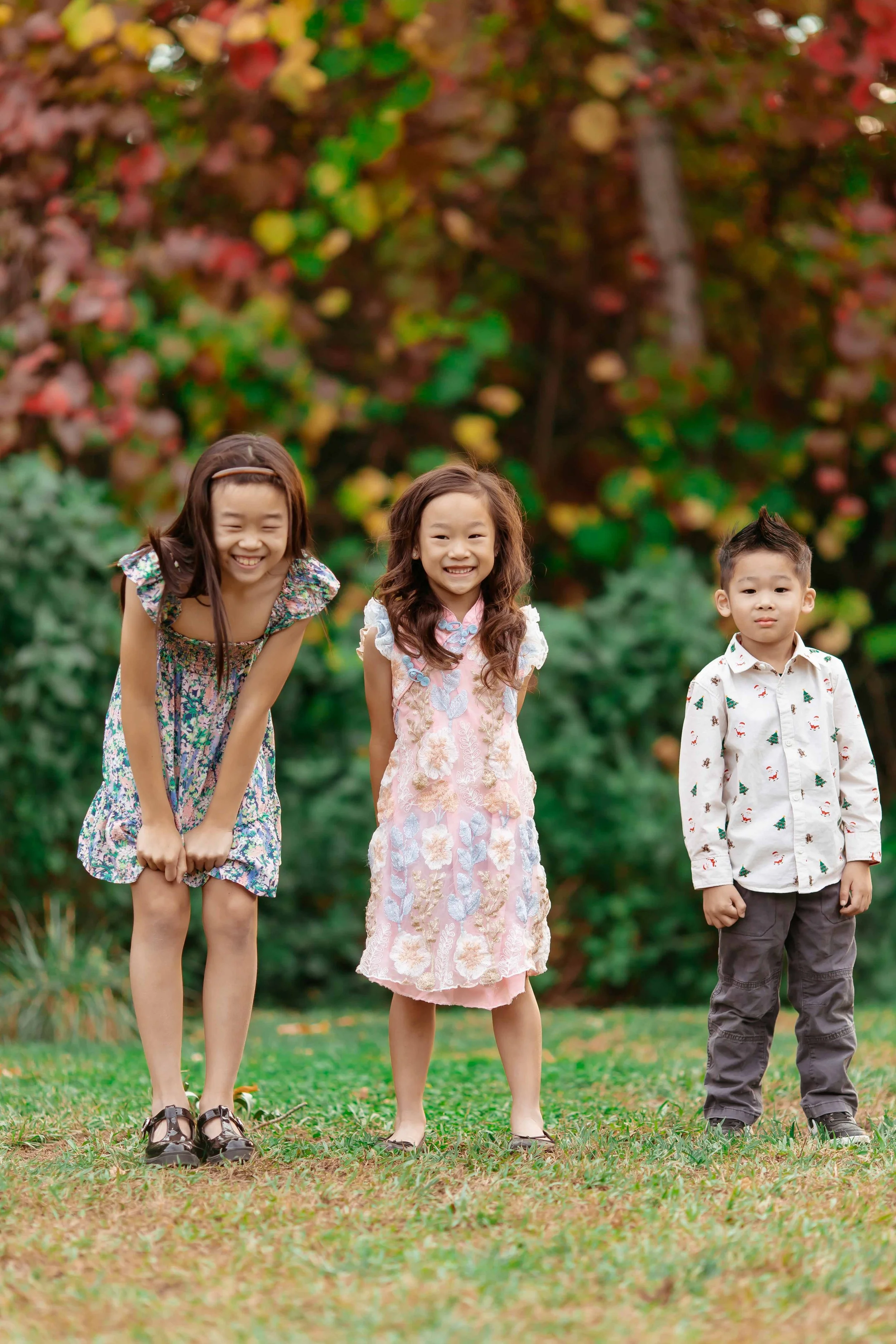 Three children standing outdoors on grass, smiling, with colorful fall foliage in the background.