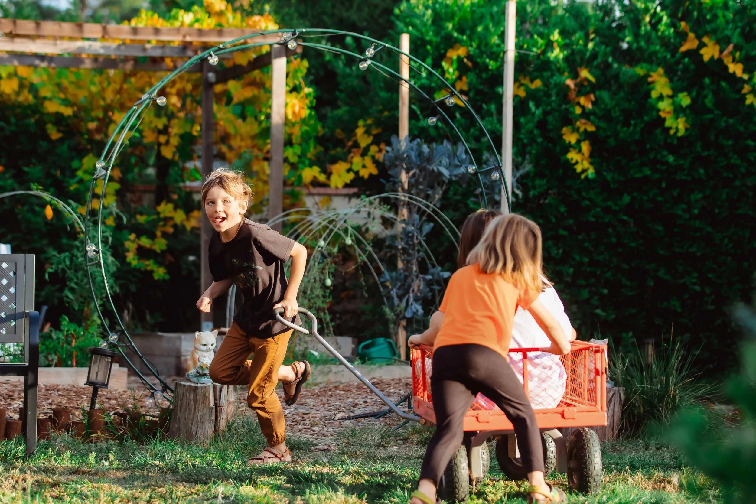 Kids playing in a garden, pulling a red wagon with two children, one in a white dress and one in an orange shirt, and a boy laughing and running.