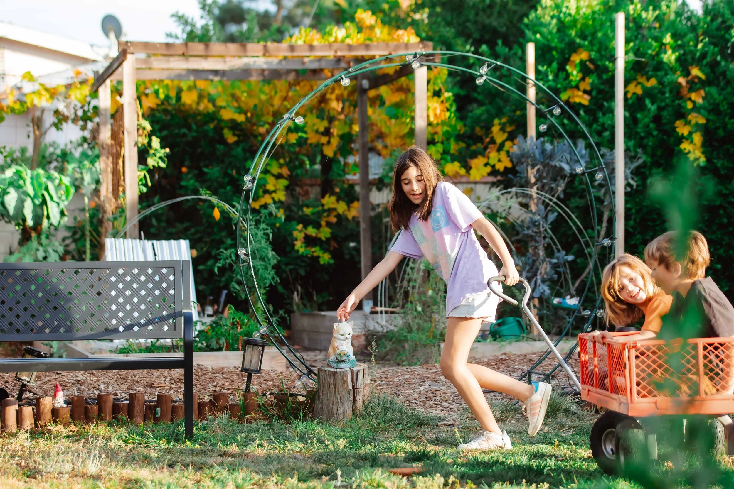 Children playing in a backyard garden with a girl reaching for a cat statue on a tree stump while two kids sit in a red wagon, surrounded by garden plants, trees, and garden structures.
