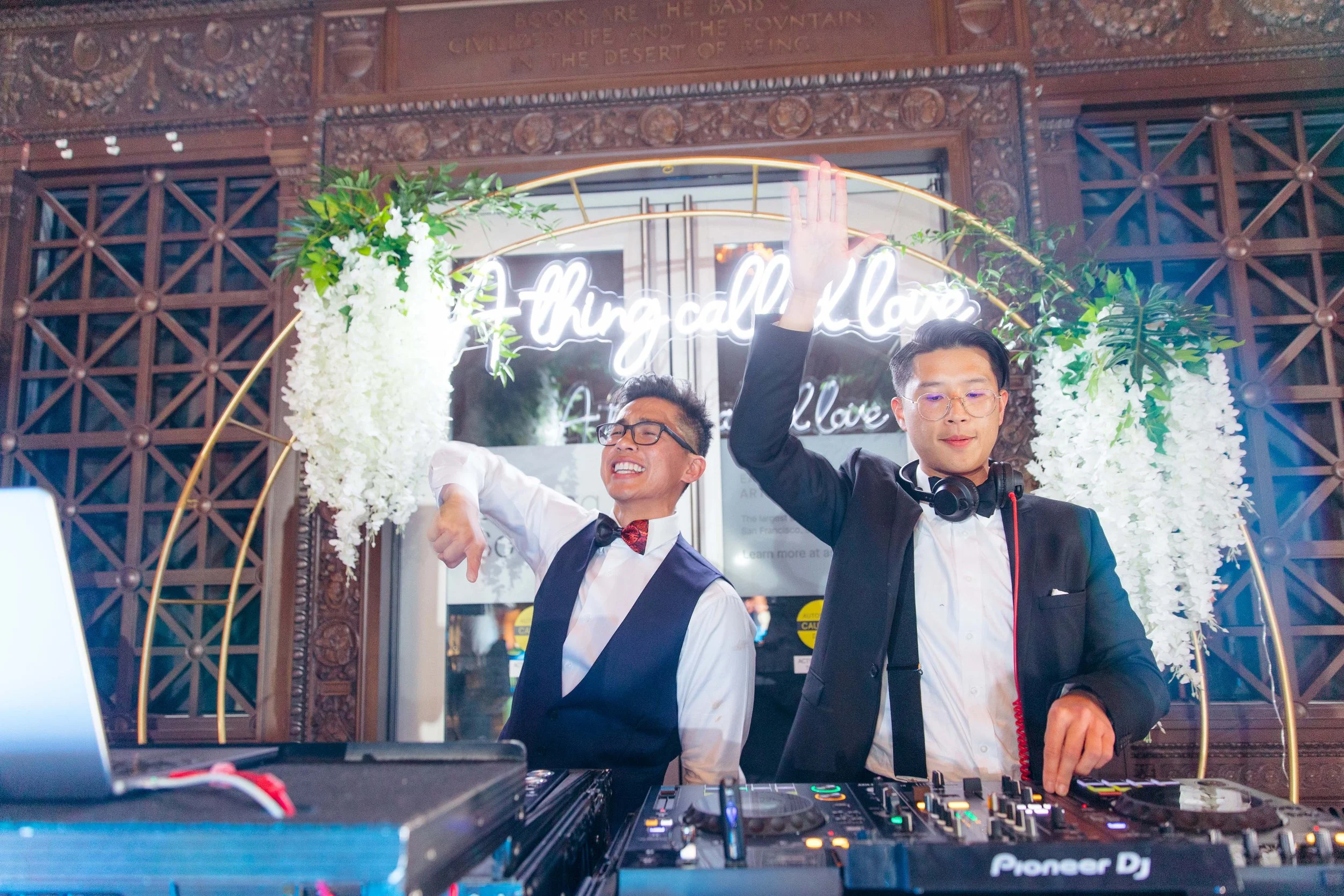 Two men in formal attire, one with glasses and a bow tie, and the other with glasses and headphones, are dancing and smiling behind DJ equipment at a celebration event with floral decorations and a neon sign that reads 'Nothing call... love' in the b