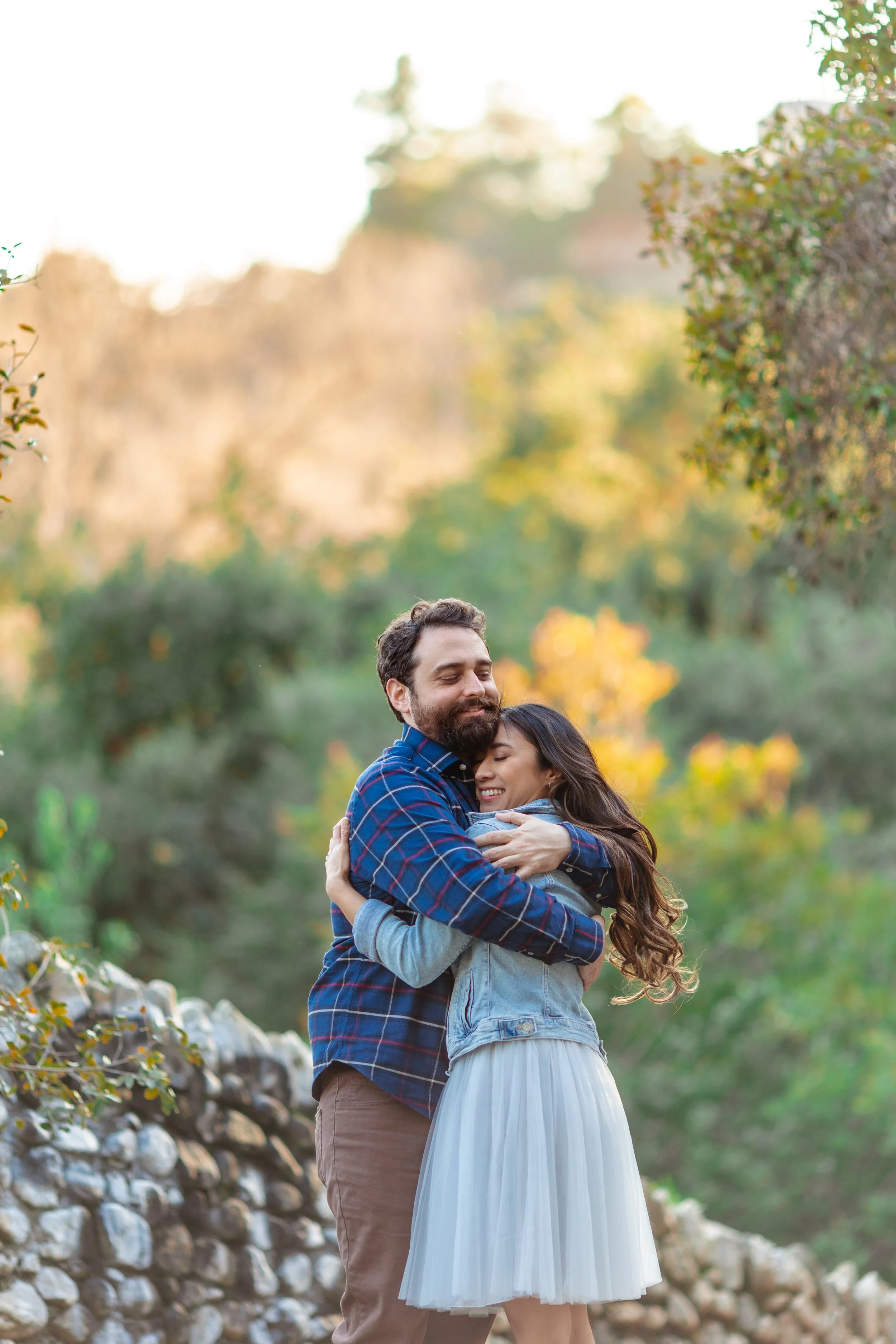 A couple hugging outdoors during autumn, with trees and a stone wall in the background.