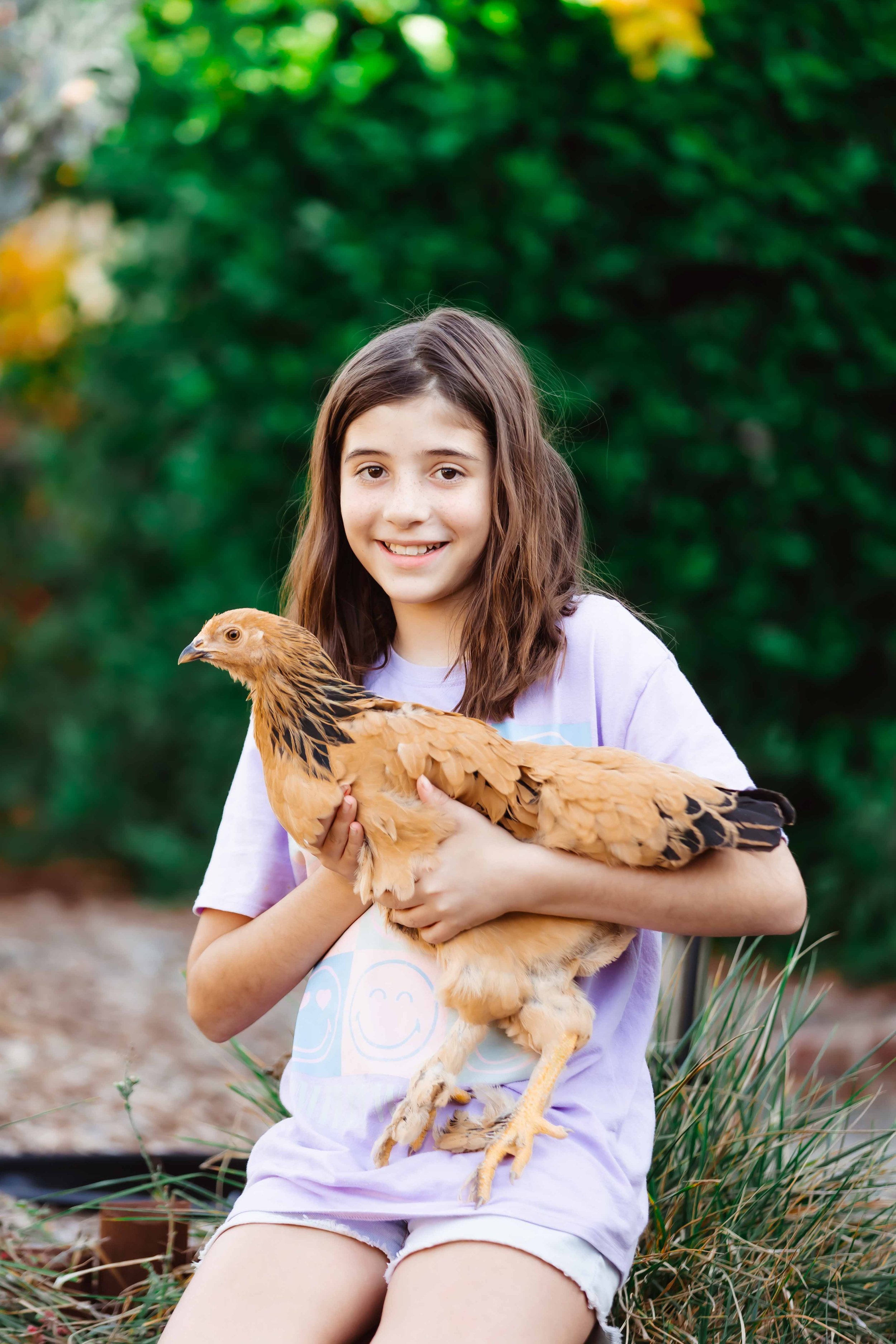 A girl with long brown hair holding a brown chicken outdoors, with green foliage in the background.