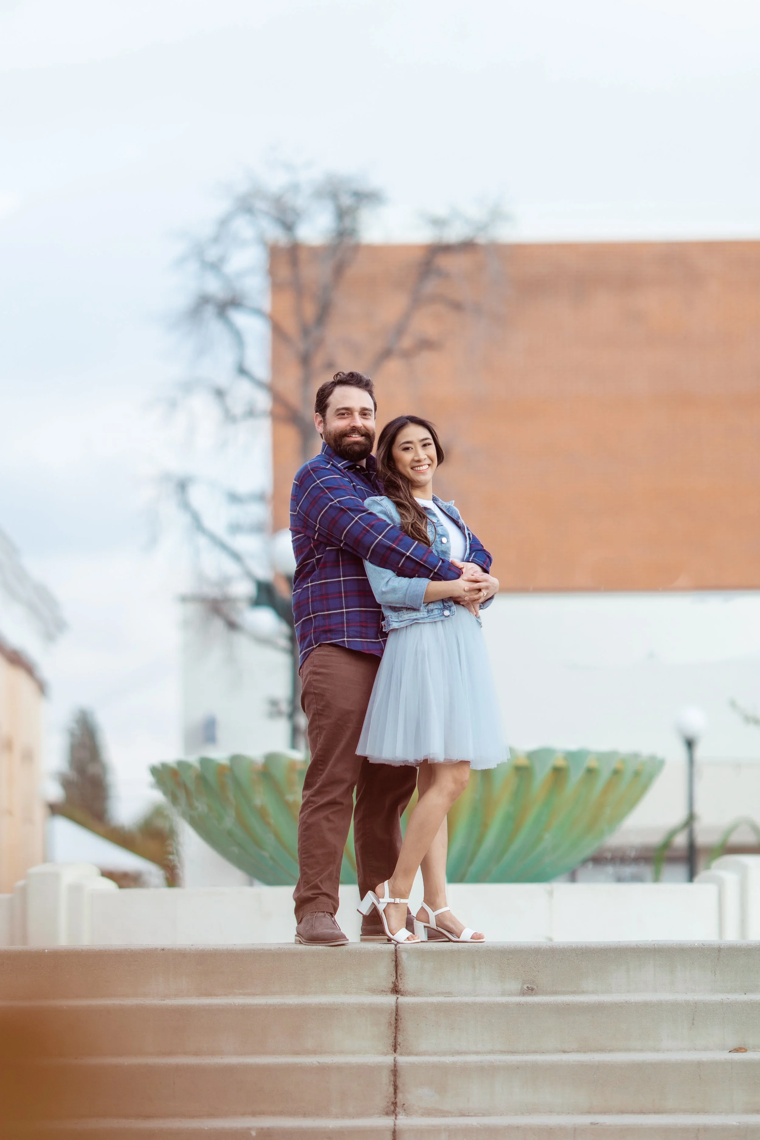 A smiling couple standing on steps in front of a large fountain, with the man having a beard and wearing a plaid shirt, and the woman in a light blue dress and white heels.