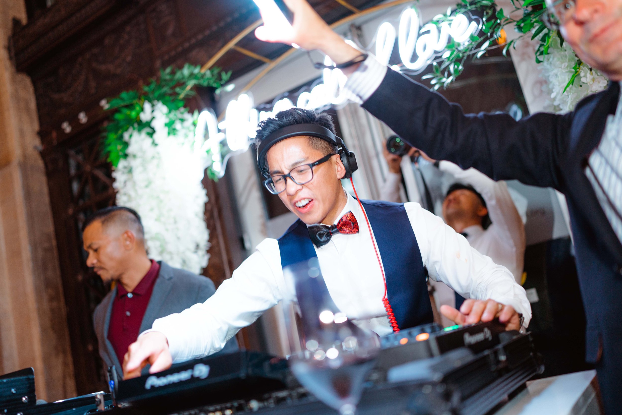 DJ in formal attire with a bow tie and glasses mixing music at an event with decorative floral background and neon sign
