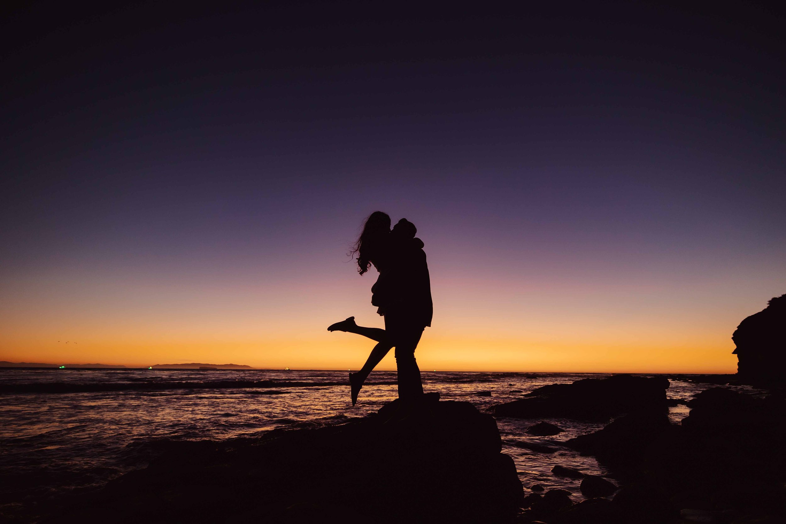 Silhouette of a couple kissing on rocks by the ocean at sunset with a colorful sky.