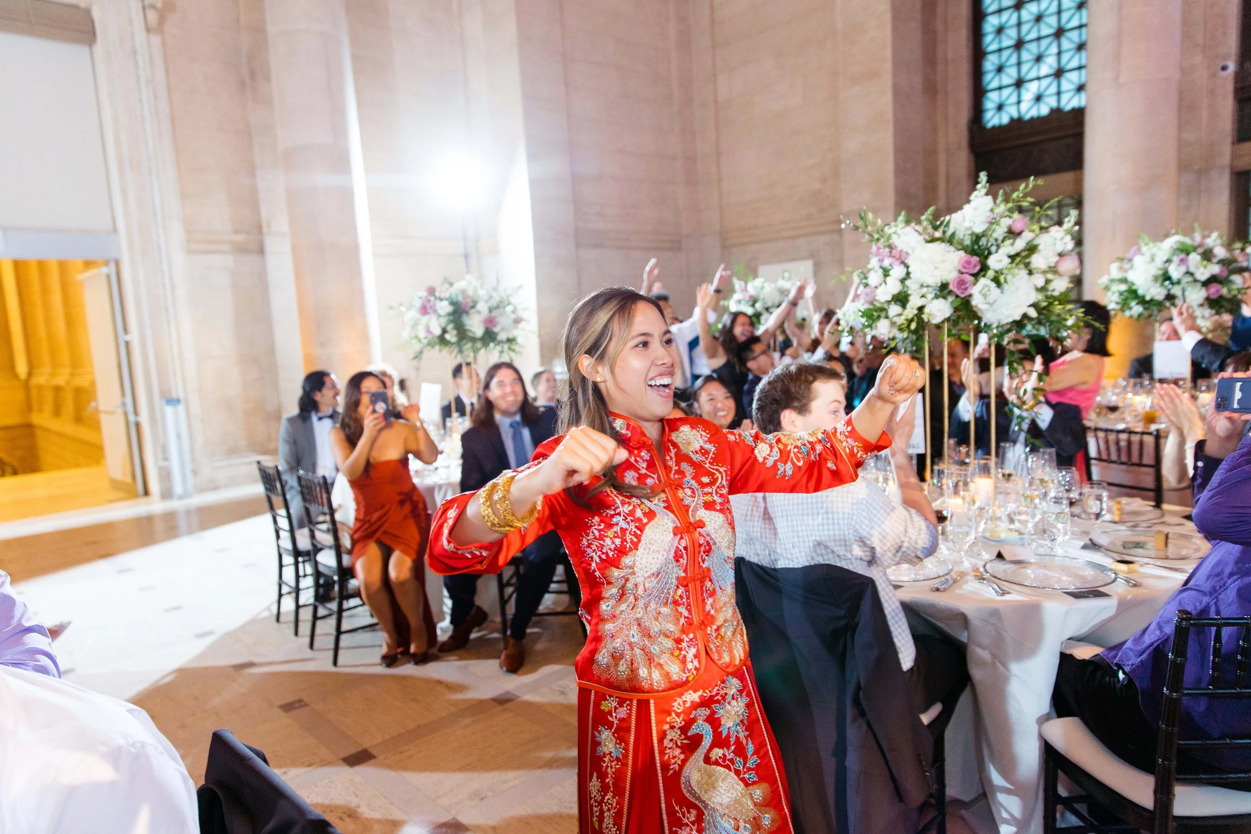 Woman in red traditional Asian dress celebrating at a formal event with guests seated at decorated tables.