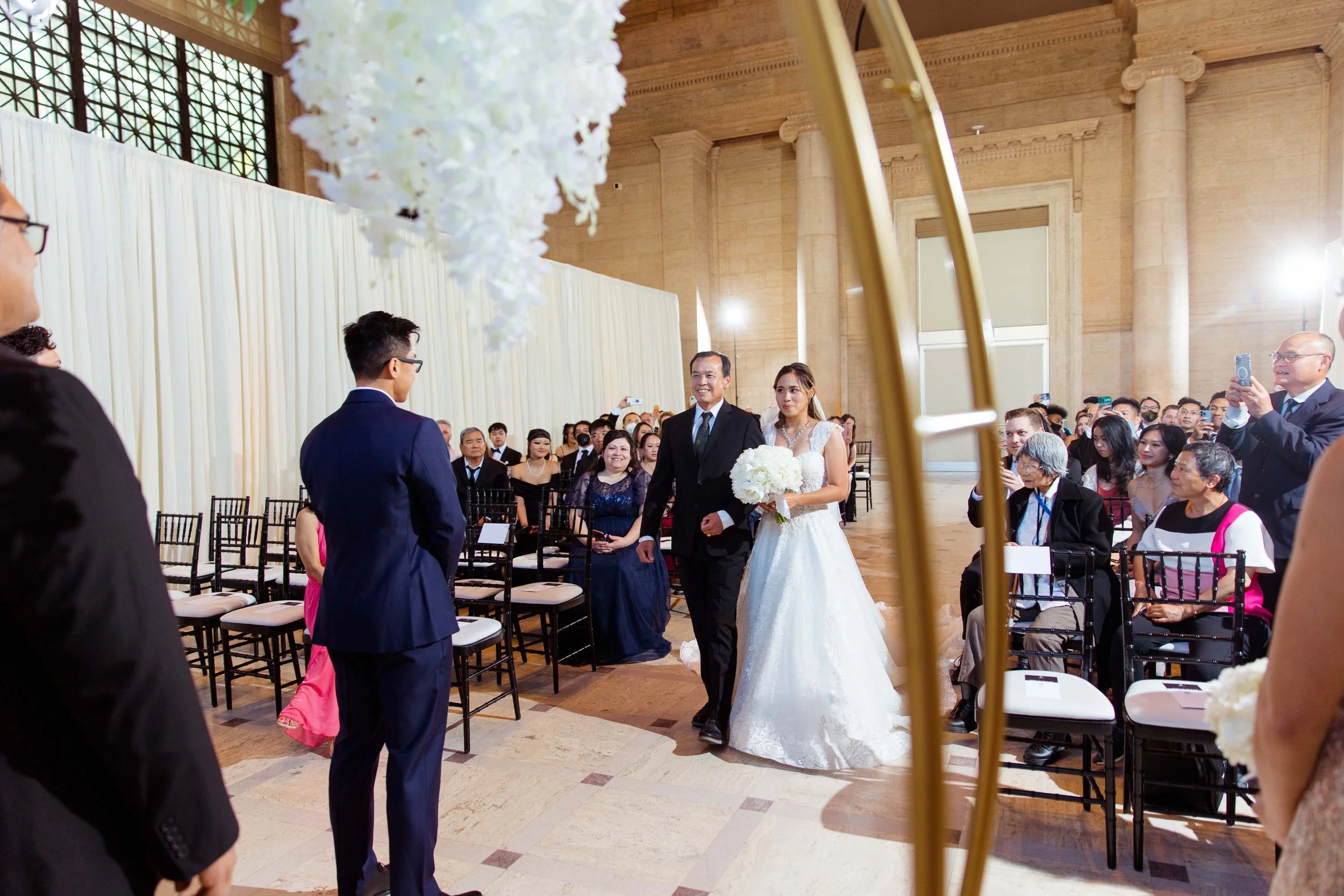 A bride walking down the aisle with her father at a wedding ceremony in a large, elegant hall filled with seated guests. The bride is in a white wedding gown holding a bouquet, and the father is in a black suit. The groom is standing at the altar in 