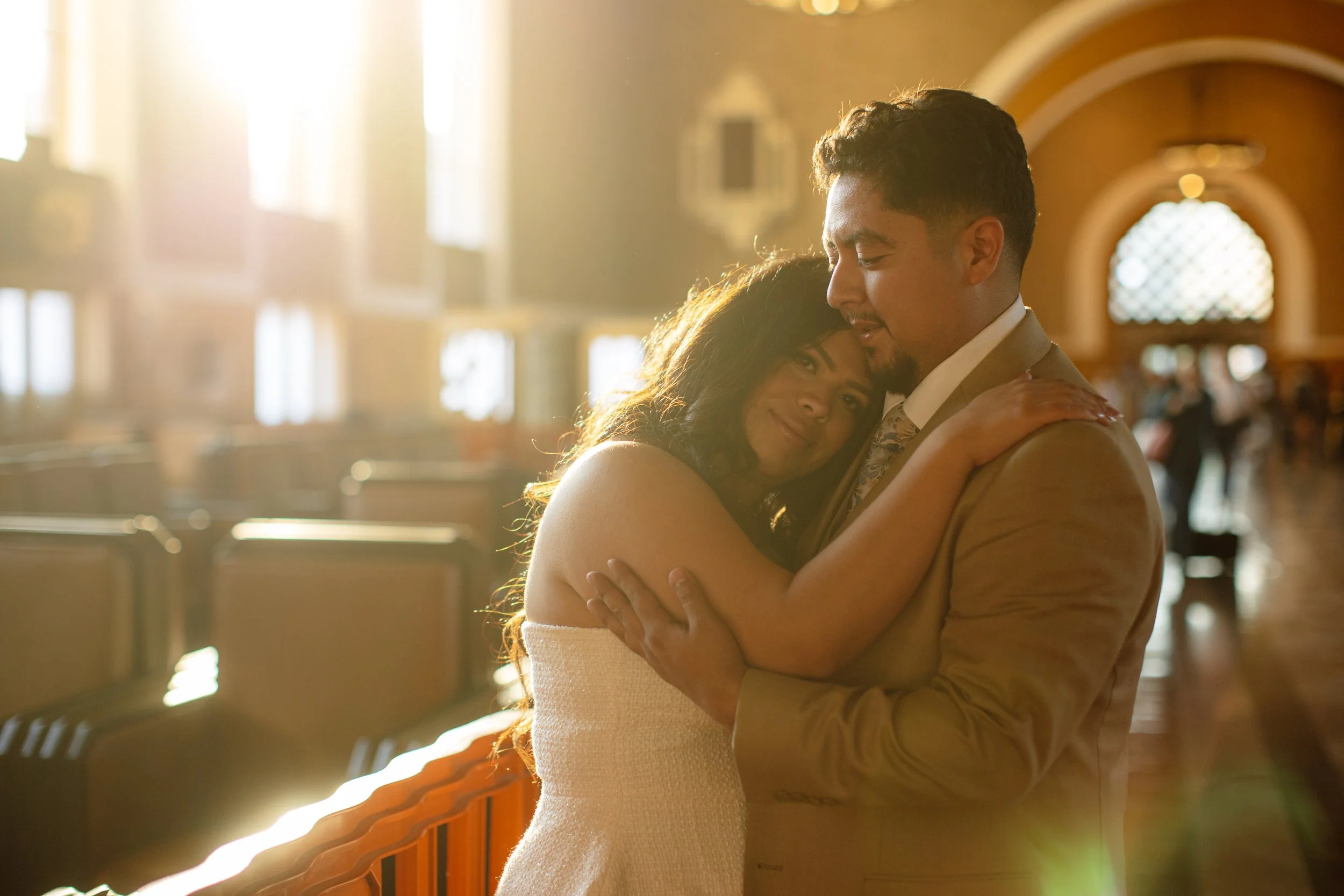 A couple hugging inside a church, with sunlight streaming through the windows in the background.