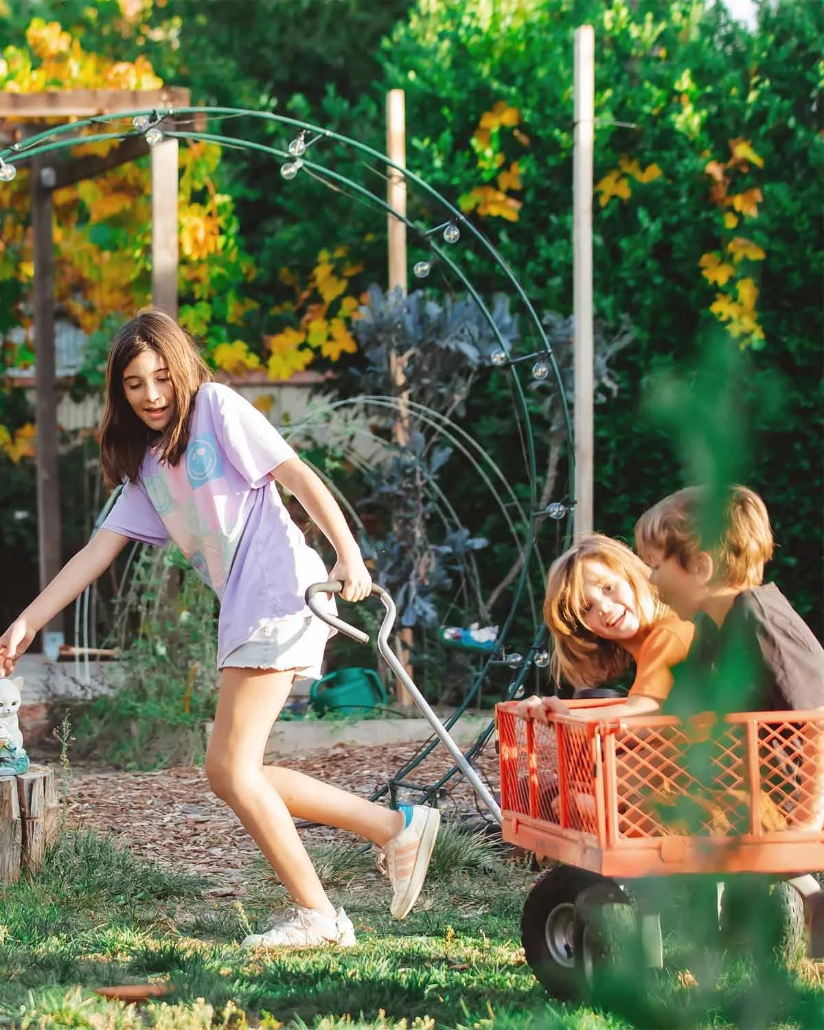 Three children playing outdoors in a garden, with one girl pulling a red wagon carrying two kids, and a girl reaching for a small figurine or object on a tree stump.