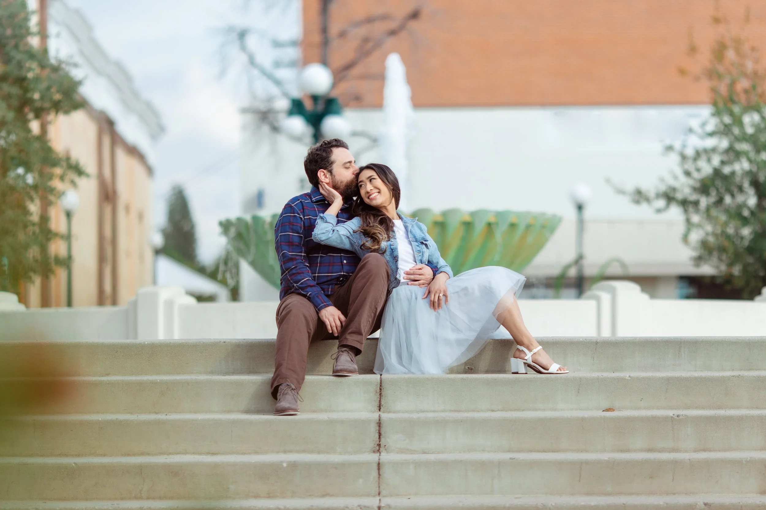 A couple sitting on steps outdoors, smiling and showing affection, with a fountain and buildings in the background.