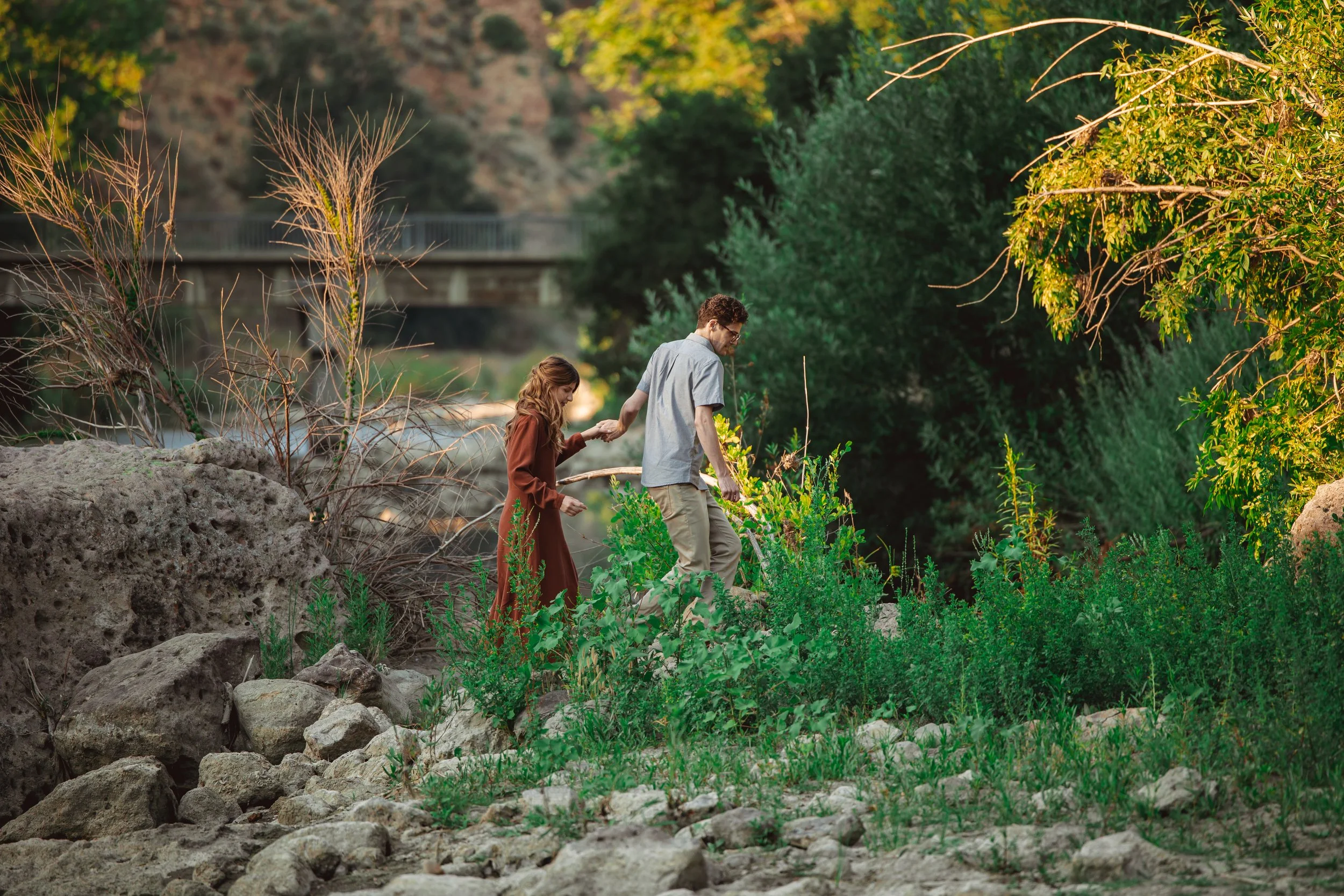 A couple hiking on rocky terrain with green bushes and trees, near a river or creek, with a bridge in the background during daytime.