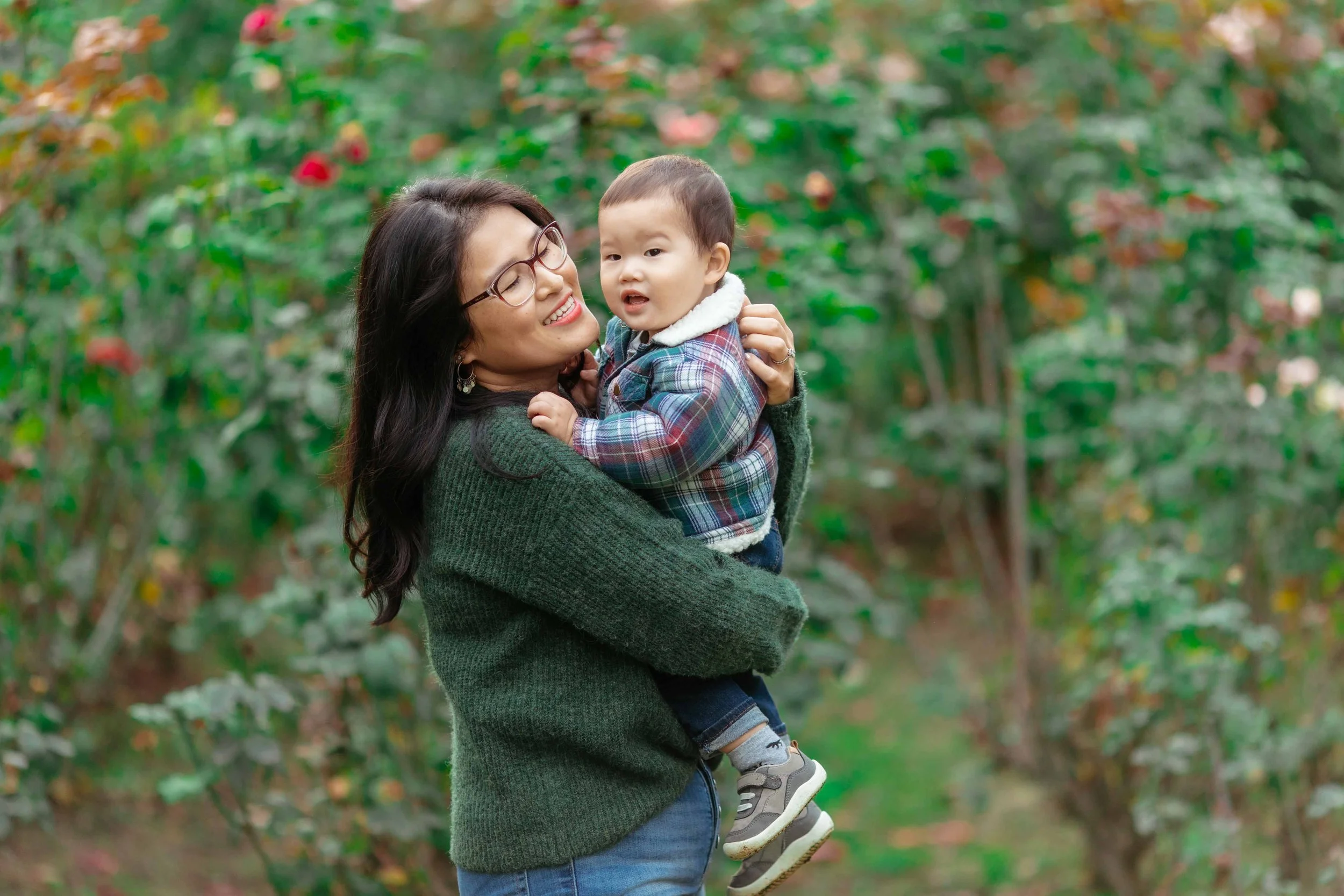 A woman with glasses and long dark hair holding a young boy in a plaid jacket and sneakers in a lush outdoor setting with greenery and trees.