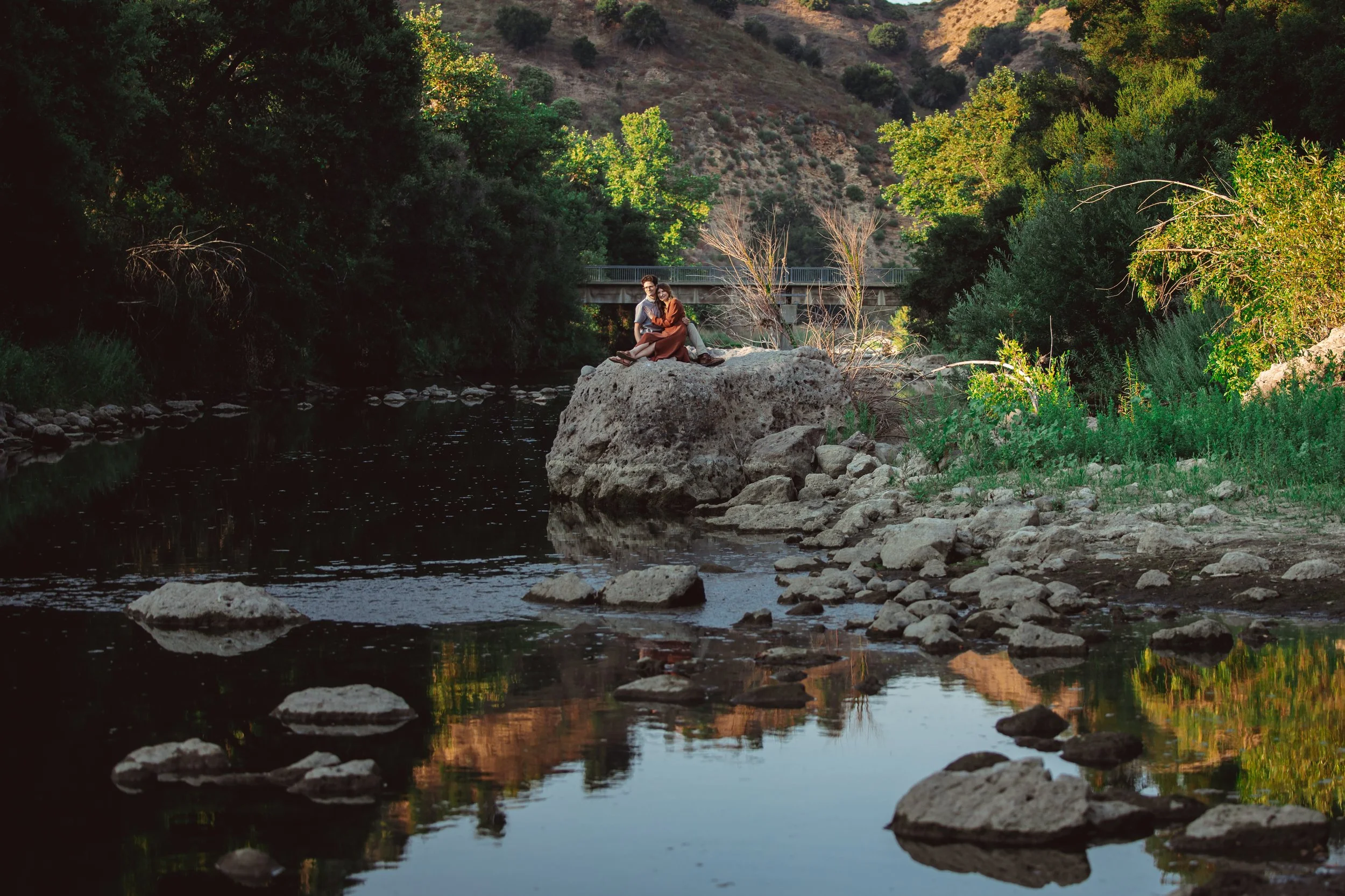 A couple sits atop a large rock in the middle of a river, surrounded by green trees and hills, with a bridge in the background.
