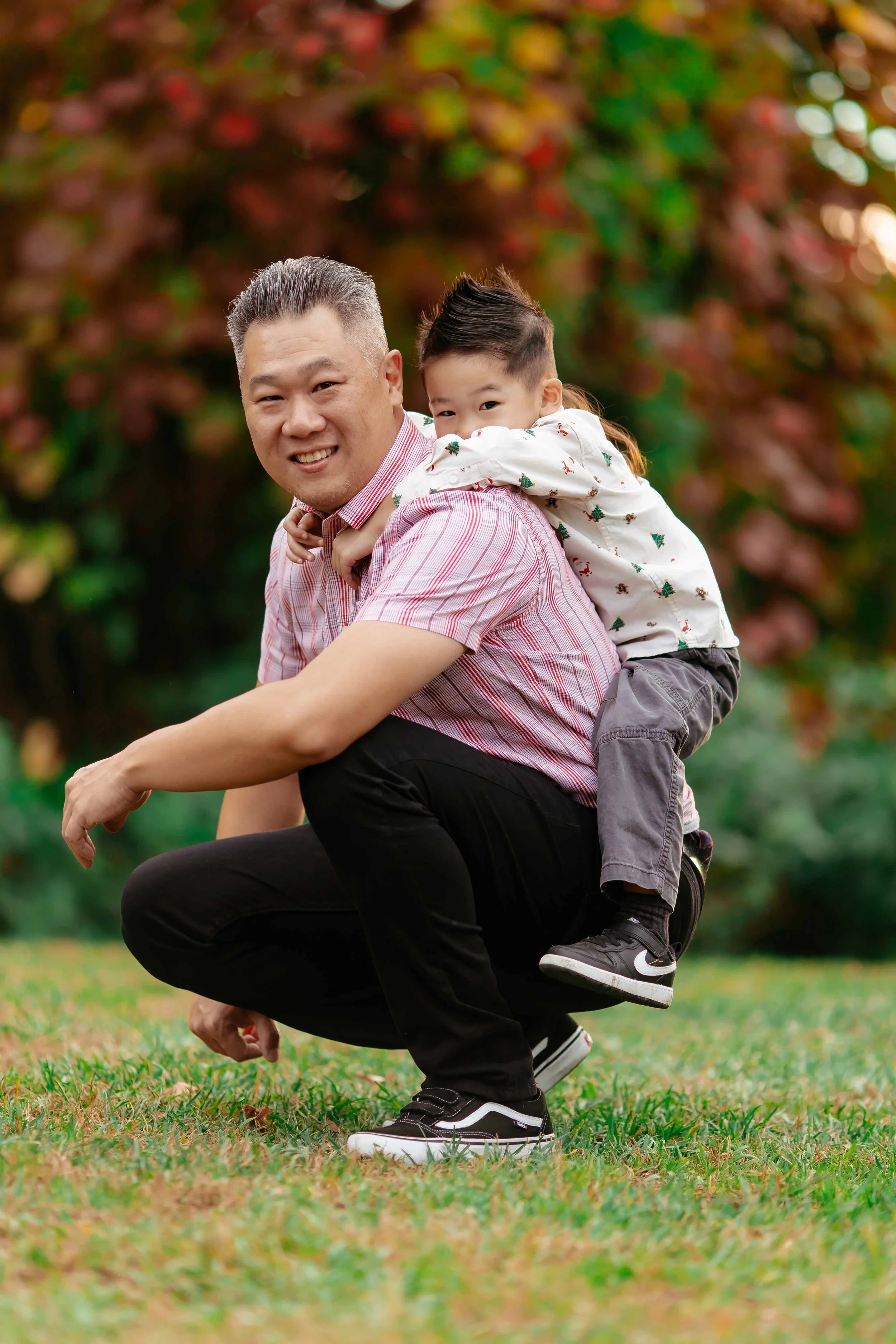 A man crouching on grass with a young boy riding on his back, both smiling. They are outdoors with colorful trees in the background.