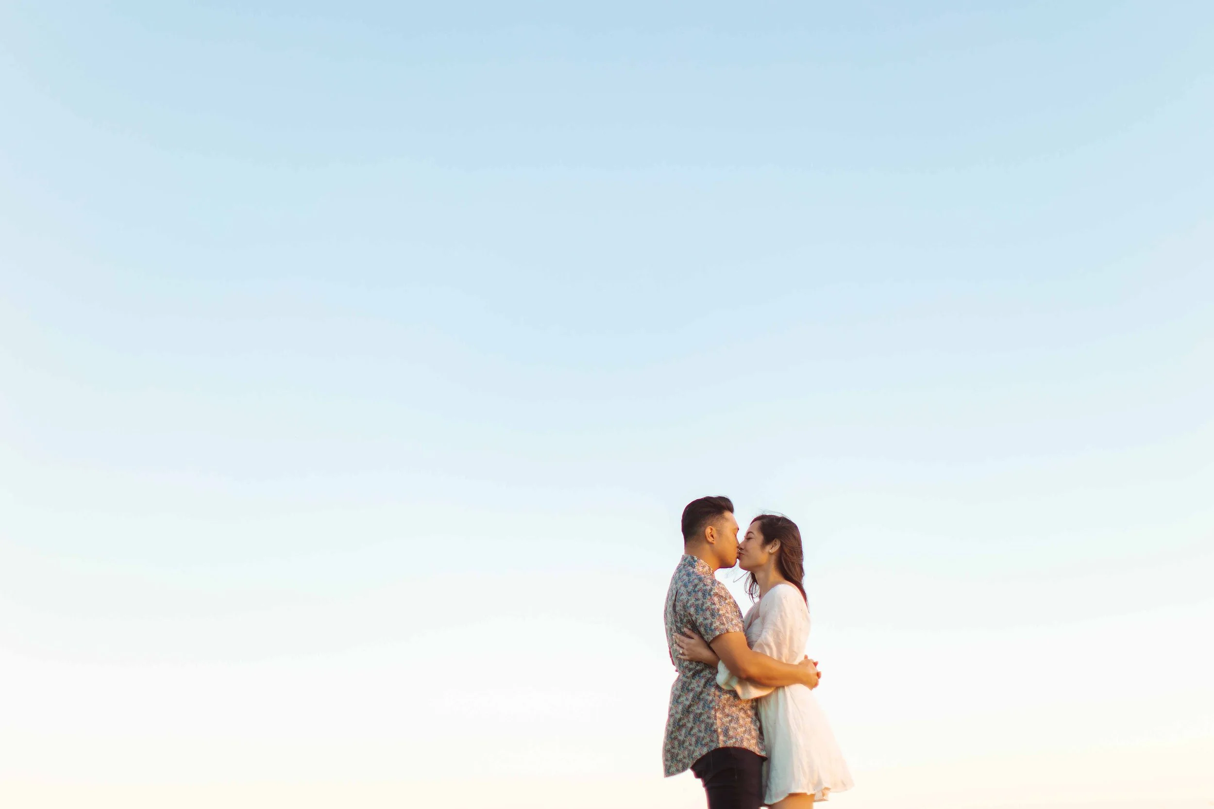 A couple is standing close together and about to kiss in an open outdoor space with a clear blue sky.