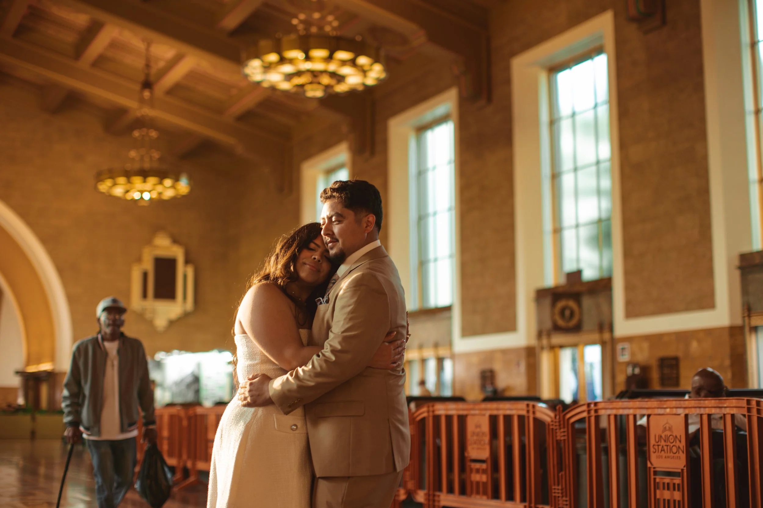 A couple embraces in a church with large windows, a high wooden ceiling, and a vintage clock, while an elderly man with glasses and a cane looks on in the background.