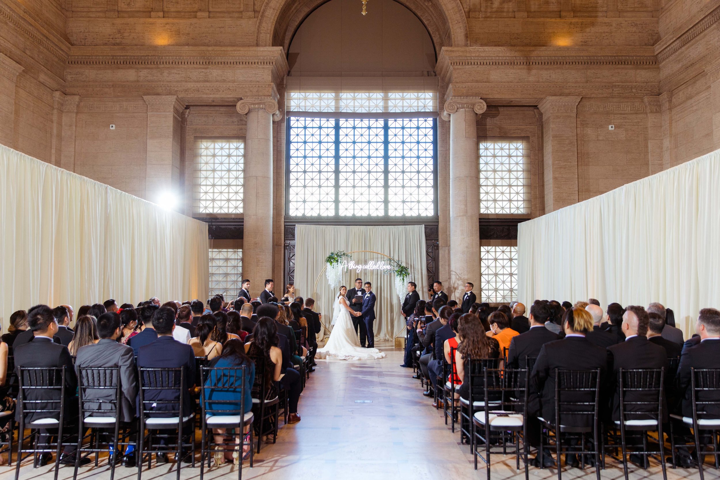 A wedding ceremony inside a grand hall with tall columns and large windows. The bride and groom stand at the altar, surrounded by attendees seated in rows. There is a decorative arch with greenery and a sign that says 'A ring called love.'