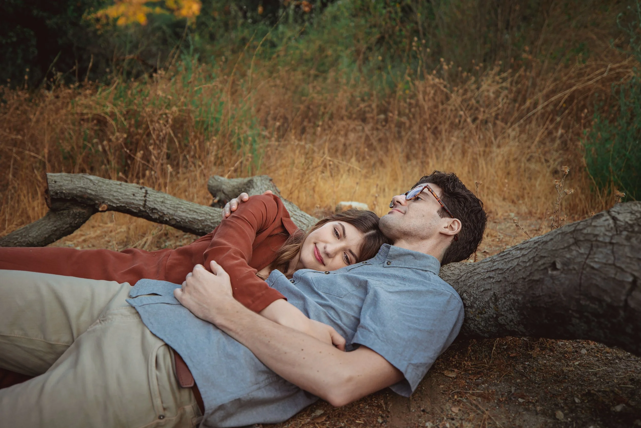 A couple lying on the ground with a fallen tree behind them, surrounded by autumn foliage.